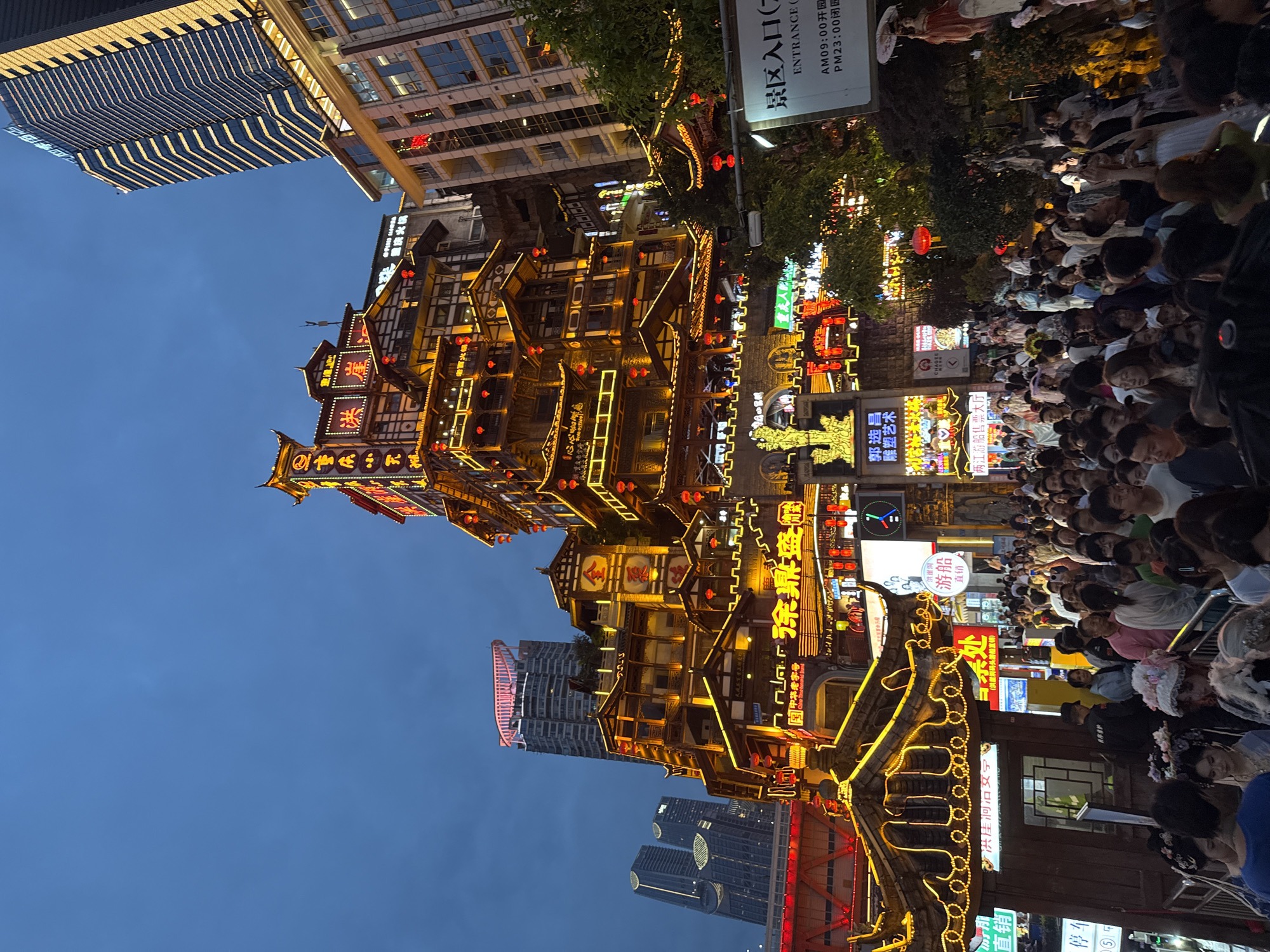 The image depicts a bustling night scene in the Yuzhong District of Chongqing, China, likely at Hongyadong. A multi-tiered, traditional Chinese architectural complex, illuminated by warm yellow lights and red lanterns, dominates the center. The building features intricate wooden structures with multiple balconies and roofs, adorned with glowing signs. In the background, modern skyscrapers with illuminated windows rise into the twilight sky. A large crowd of people is visible, walking and standing along a pathway in front of the complex. Some individuals appear to be tourists, while others might be residents. The atmosphere is lively, with many people holding up phones to take pictures. A sign with Chinese characters and "ENTRANCE" in English is partially visible on the right, indicating it's a popular tourist destination. The sky is a deep blue, suggesting it's either dusk or early evening.