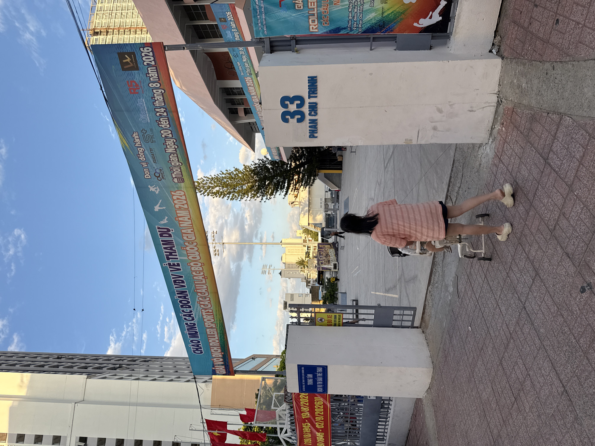 An exterior shot captures a wide concrete entrance to a roller sports facility in Nha Trang, Vietnam. A large banner spanning across the entrance reads in Vietnamese, "CHAO MUNG CAC DOAN VDV VE THAM DU GIAI VO DICH ROLLER SPORTS CAC CAU LAC BO QUOC GIA NAM 2026" which translates to "Welcome to the VDV delegations to participate in the National Club Roller Sports Championship 2026." Below this, smaller text indicates the event dates as "Ngay 20 den 24 tháng 8 năm 2026" or "August 20-24, 2026." On the right side of the entrance, a white pillar displays the blue text "33 PHAN CHU TRINH," indicating the street address. A young woman wearing a pink striped t-shirt and dark shorts walks away from the camera, pushing a stroller. The sky above is blue with scattered white clouds. In the background, buildings, a tall pine tree, a basketball hoop, and streetlights are visible.