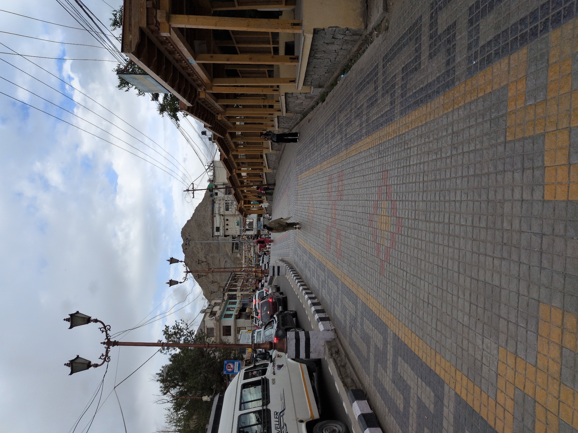 The image depicts a street scene in Leh, India, likely a market or main thoroughfare, characterized by a paved walkway with a distinctive pattern of grey, yellow, and dark grey tiles. The walkway is bordered by a road with a black and white striped curb, where a white minivan and several cars are parked or in traffic. On the right side of the street, a covered structure with wooden pillars and a sloped roof runs alongside the walkway, resembling a market stall or shopfront. Several people are visible: a woman in a traditional garment walks on the tiled path, another woman stands near the wooden stalls, and others are seen in the distance near the parked vehicles and buildings. The background features multi-story buildings with windows and balconies, typical of urban architecture in the region. A large, barren mountain looms in the distance under a cloudy sky. The presence of street lamps, power lines, and a "No Parking" sign further indicate an urban setting. The overall impression is that of a bustling street in Leh, with its unique architectural style and mountainous backdrop.