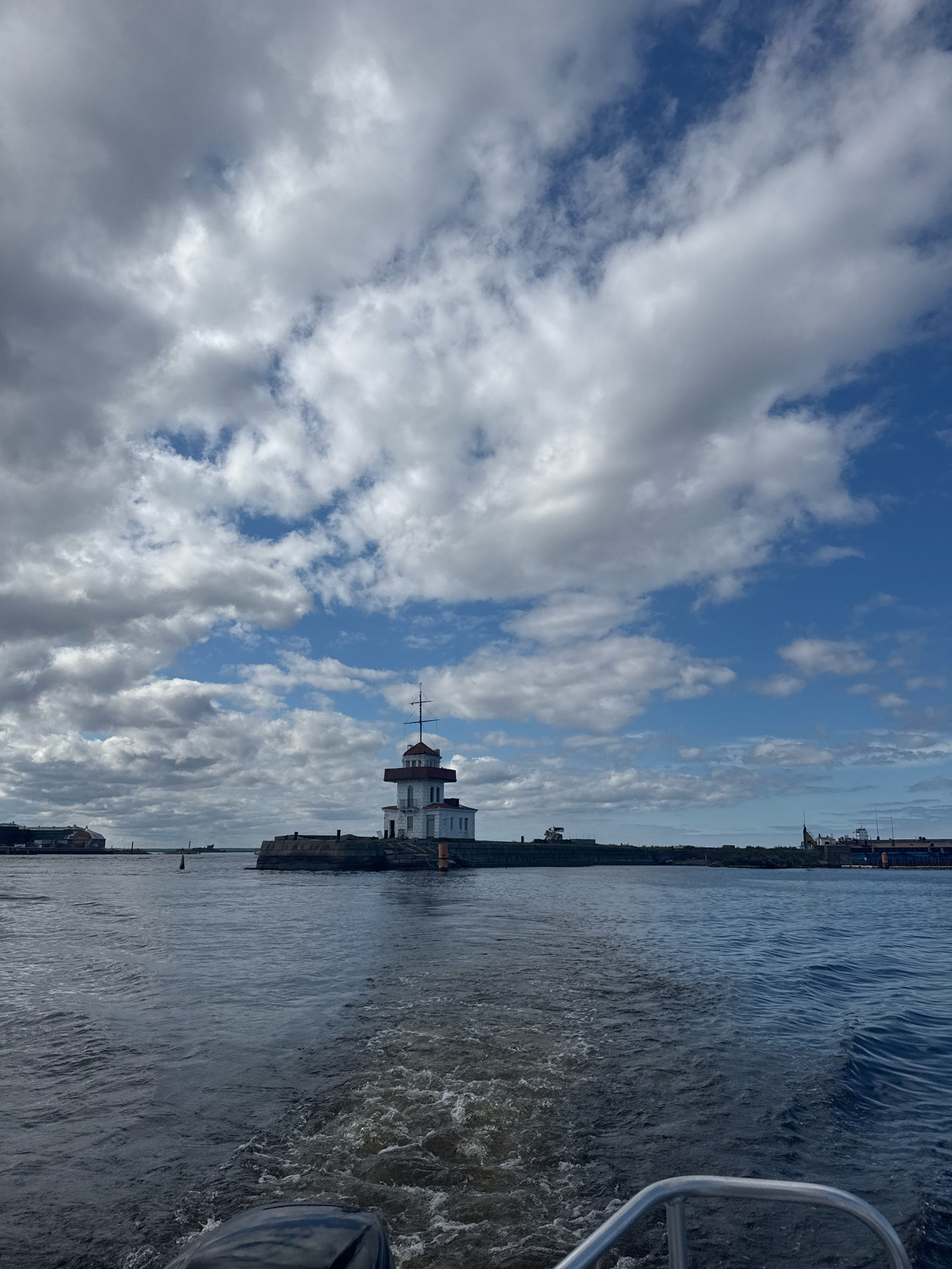 A wide-angle, eye-level shot captures a lighthouse situated on a stone pier in the waters of Kronstadt, Russia. The lighthouse, painted white with a red roof and a lantern room, stands prominently against a backdrop of a dynamic sky filled with a mix of white and grey clouds. The choppy blue water in the foreground suggests the perspective is from a moving boat, with a wake visible behind it. To the left, a long, low building with a red roof is visible on the shore, and to the right, industrial structures and cranes are present. The overall scene conveys a nautical, perhaps historical, atmosphere.