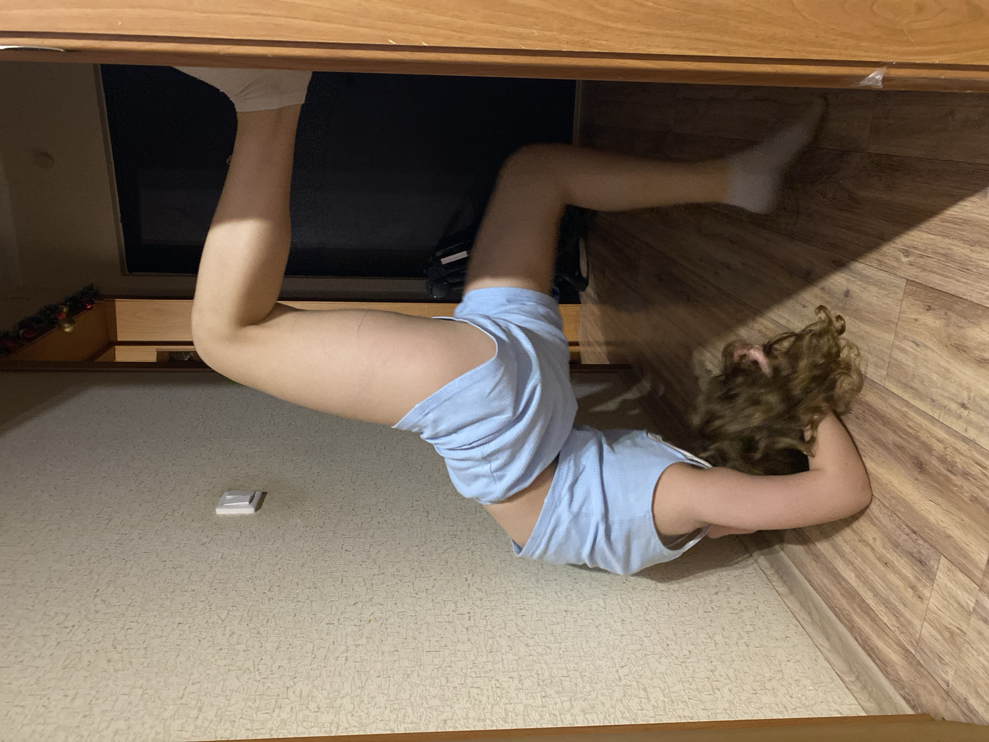A child is positioned under a wooden structure, possibly a cabinet or bed. The child wears light blue shorts and a matching top, with white socks on their feet. One leg is extended upwards and into the dark space beneath the structure, while the other is bent at the knee and extends towards the corner of the room. The child's head rests on the floor against the wall, with curly hair visible. The floor is covered in patterned linoleum. A small, rectangular object is on the floor near the center of the frame. The lighting in the room is dim. The location appears to be an indoor residential space in an unspecified city.