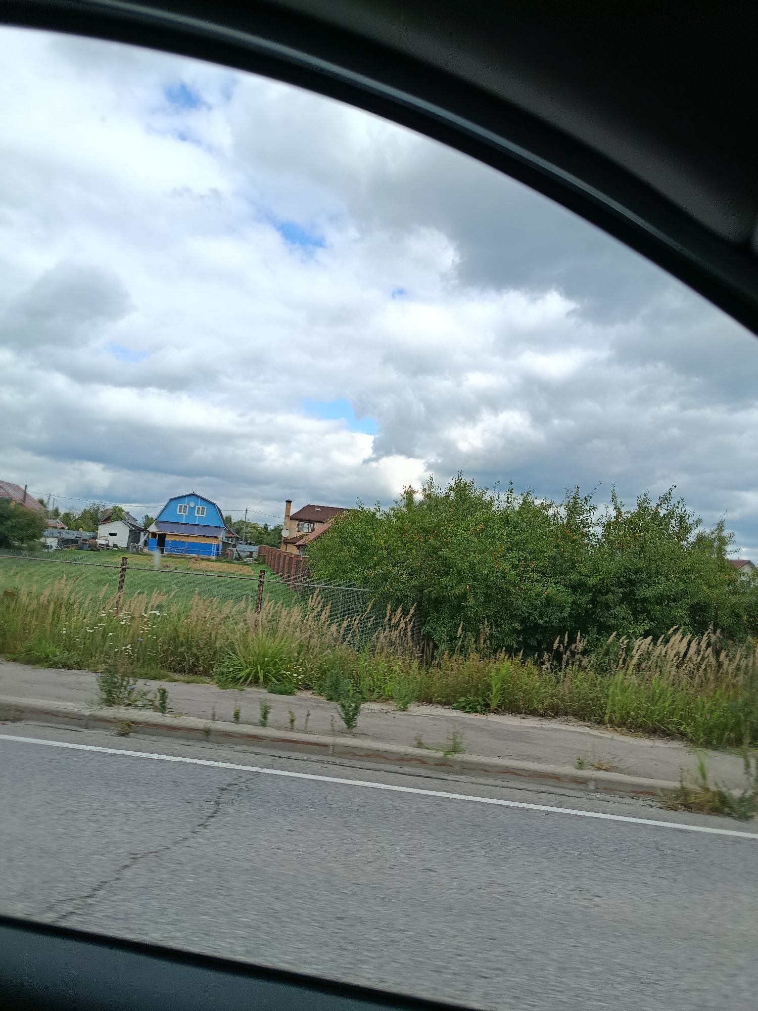 The image shows a rural scene from the perspective of a vehicle, likely a car. In the foreground, a paved road with a white line is visible, with a strip of vegetation and a fence beyond it. Behind the fence, there are several houses, including a distinct blue and yellow house with white-framed windows. Tall green trees and bushes are prominent in the midground. The sky is partially cloudy, with patches of blue sky visible between the white and grey clouds. The overall impression is that of a suburban or rural residential area under an overcast sky.
