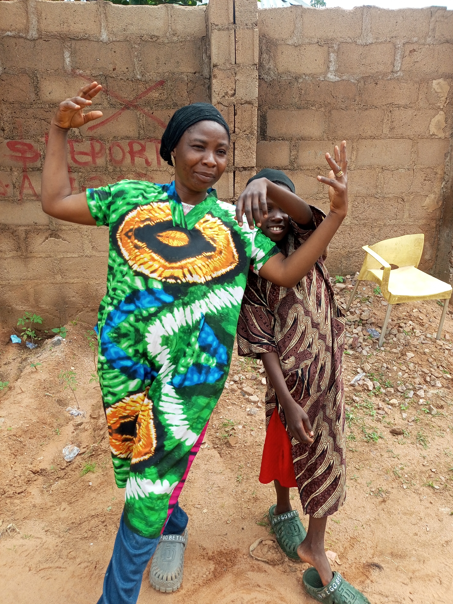 In Tempure, Nigeria, two individuals, likely a mother and child, pose for a photograph in front of a rough, tan-colored brick wall. The wall has red spray-painted markings that appear to be letters and an "X". The ground is sandy with scattered rocks and small patches of greenery.
The taller individual, on the left, is a woman wearing a black headscarf and a vibrantly colored dress with green, blue, yellow, and black patterns. She is flexing her left arm, with her hand open and fingers spread. Her right arm is bent at the elbow, with her hand raised and fingers curled. She is looking at the camera with a slight smile.
The shorter individual, to the right, is a child wearing a dark headscarf and a patterned brown dress over a red garment. The child is also striking a pose, with their left arm bent across their face and their right hand raised, fingers spread. The child is smiling broadly at the camera. Both individuals are wearing open-toed sandals.
In the background, to the right, a bright yellow plastic chair sits on the uneven ground. The overall scene suggests an informal outdoor setting, possibly a residential area in Tempure, Nigeria.