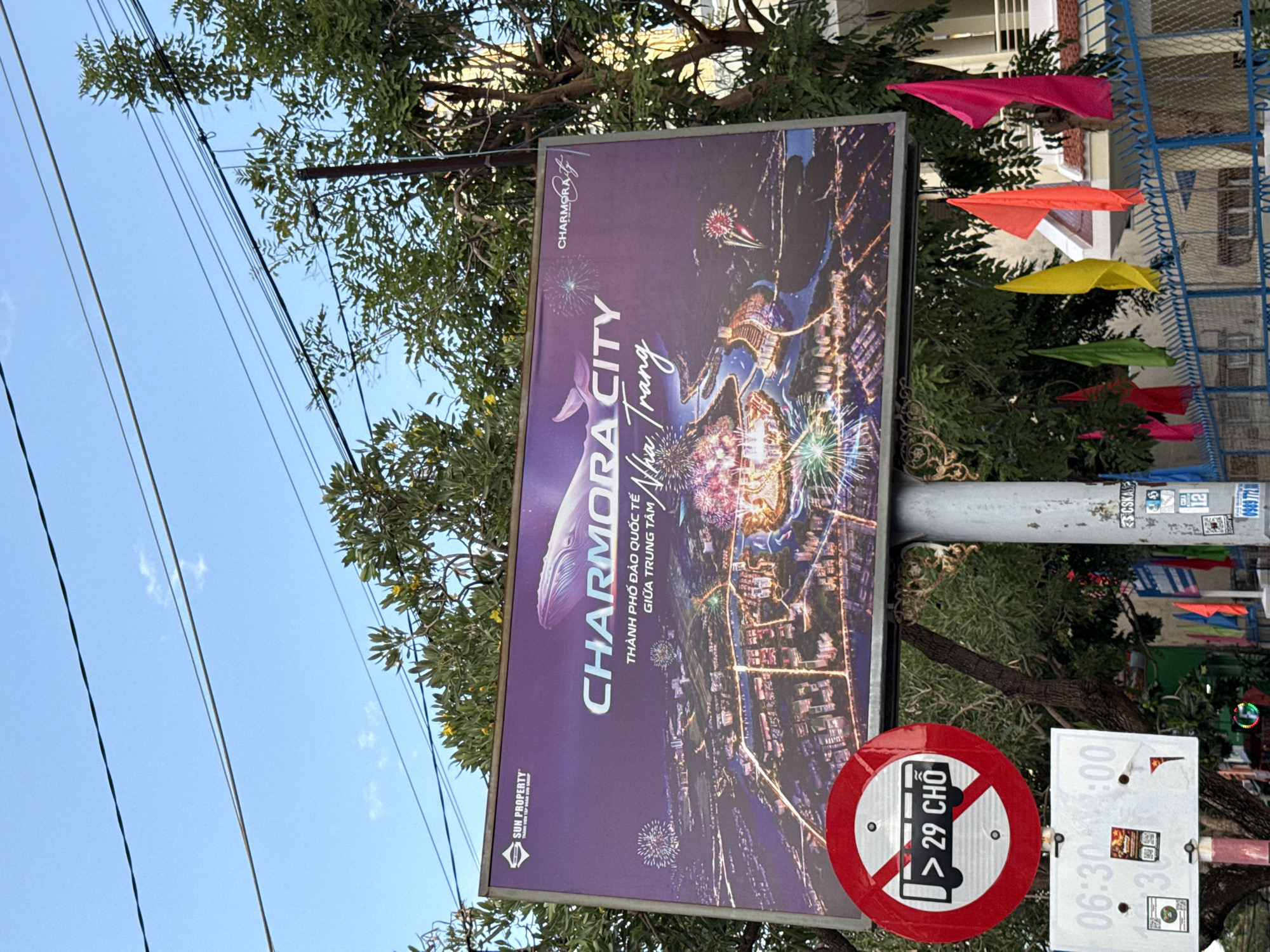 The image shows a billboard advertising "CHARMORA CITY" in Nha Trang, Vietnam. The billboard displays an aerial view of a city illuminated by fireworks and a large whale graphic. Below the billboard, there is a traffic sign indicating "NO BUSES OVER 29 SEATS". A white sign with black text displays "06:30 - 11:00". The scene is set outdoors with trees and a blue fence in the background. The sky is clear with some visible power lines.