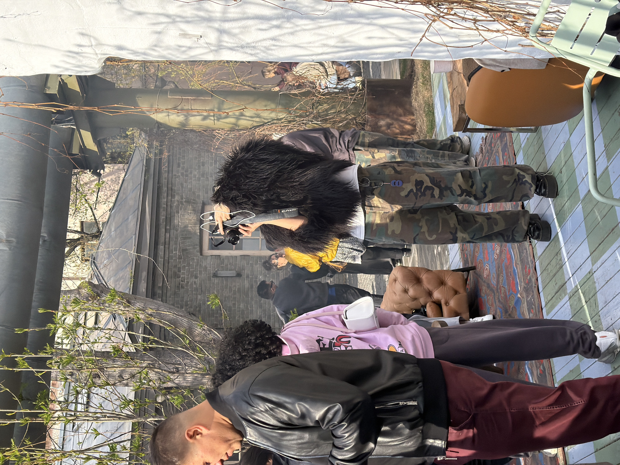 The image shows a group of people in an outdoor setting. The background includes a brick building and metal pipes, suggesting an urban environment, possibly in Moscow, Russia. Several individuals are visible, dressed in contemporary clothing. One person is wearing camouflage pants and a fur-like collar, holding a camera, possibly taking photographs. Others are wearing casual attire, including a pink hoodie and a leather jacket. There are chairs and a rug visible on a wooden surface, indicating a staged or arranged setting, possibly for a gathering or an event. The presence of branches and nascent greenery suggests the season is spring.