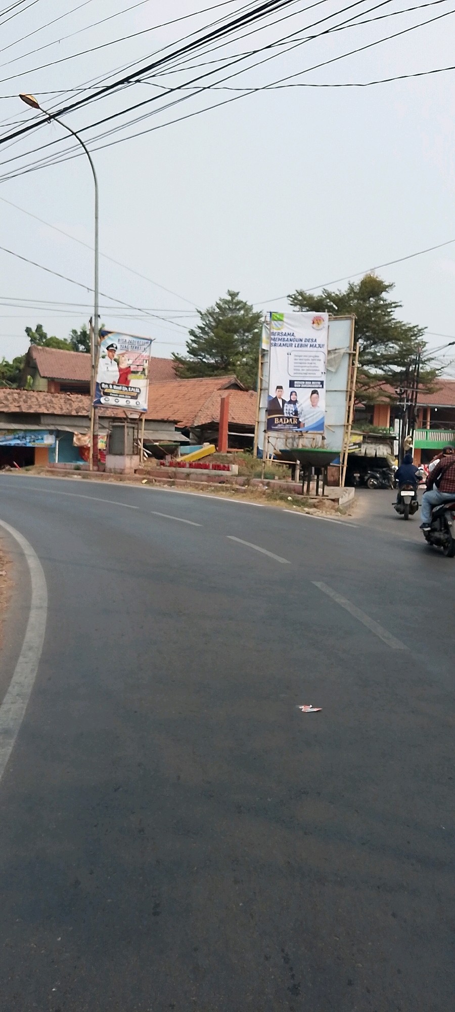 A street scene in Satriamekar, Indonesia, features a two-lane asphalt road with a white lane divider. The road curves to the left, with a concrete curb on the left edge and a low brick wall on the right. Two billboards are visible on the right side of the road. The left billboard has images of two people and text in Indonesian. The right billboard is larger and also has images of people and Indonesian text. Behind the billboards, buildings with red tile roofs and some trees are present. A street light with a curved pole is on the left side of the image, with numerous overhead wires. Several motorcycles are on the road. On the right side of the road, two individuals are riding on a motorcycle. Further down the road, another motorcycle with a rider is visible. On the road surface itself, a small piece of red and white paper is on the asphalt. The sky is overcast.