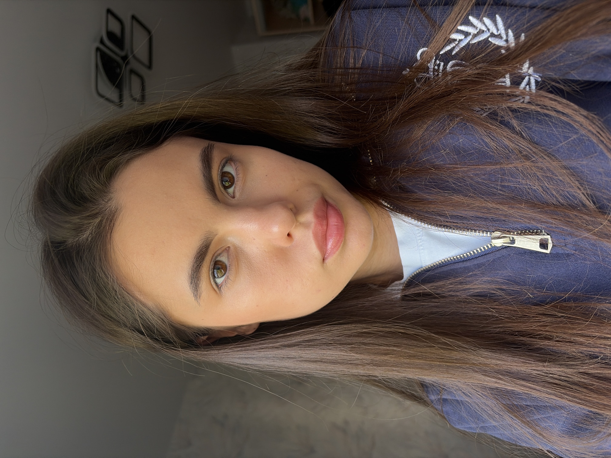 A close-up portrait shows a woman with long, straight brown hair. She has fair skin, light brown eyes, and light pink lips. She is wearing a blue zip-up jacket over a white top. The zipper of the jacket is visible. In the background, there is a blurred object on the wall. The location, Chaikovsky, Russia, cannot be independently verified from this image alone.