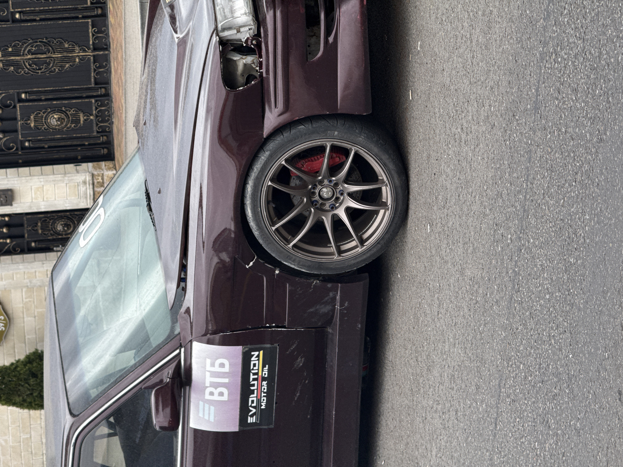 The image shows a damaged, maroon-colored car parked on an asphalt road. The car has a missing headlight and visible damage to the front fender. A sticker with "BTB" and "EVOLUTION MOTOR OIL" is on the driver's side door. The license plate is not visible. Based on the signage "59/3" and architectural style in the background, the location may be the city of Sochi, Russia. The scene does not depict any people, actions, or interactions. The environment appears to be an urban street.