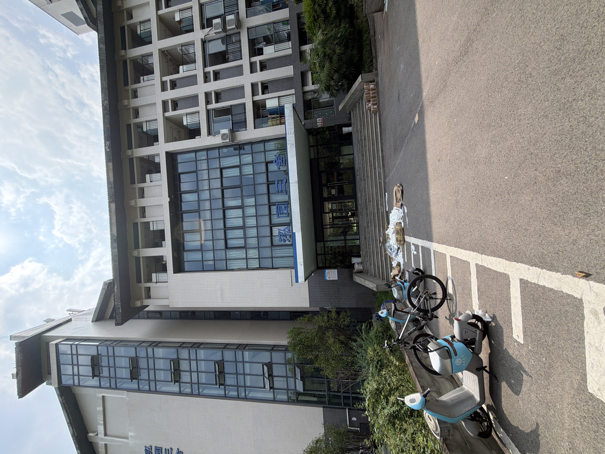 The image depicts the exterior of a modern, multi-story building, likely an institutional or residential complex, situated in Jiangjin, China. The building features a prominent glass facade on the lower floors, with multiple windows and balconies on the upper levels. Several bicycles and electric scooters are parked in front of the building, indicating a mode of transportation commonly used in the area. The scene is set against a backdrop of a bright, partly cloudy sky, suggesting daytime. Lush green trees and bushes surround the building, contributing to the urban landscape. A paved area in front of the building serves as a parking space for the vehicles and leads to a set of stairs ascending to the building's entrance. Signs with Chinese characters are visible on the building, offering insights into its function or naming. The overall impression is that of a functional, contemporary structure within a well-maintained environment.