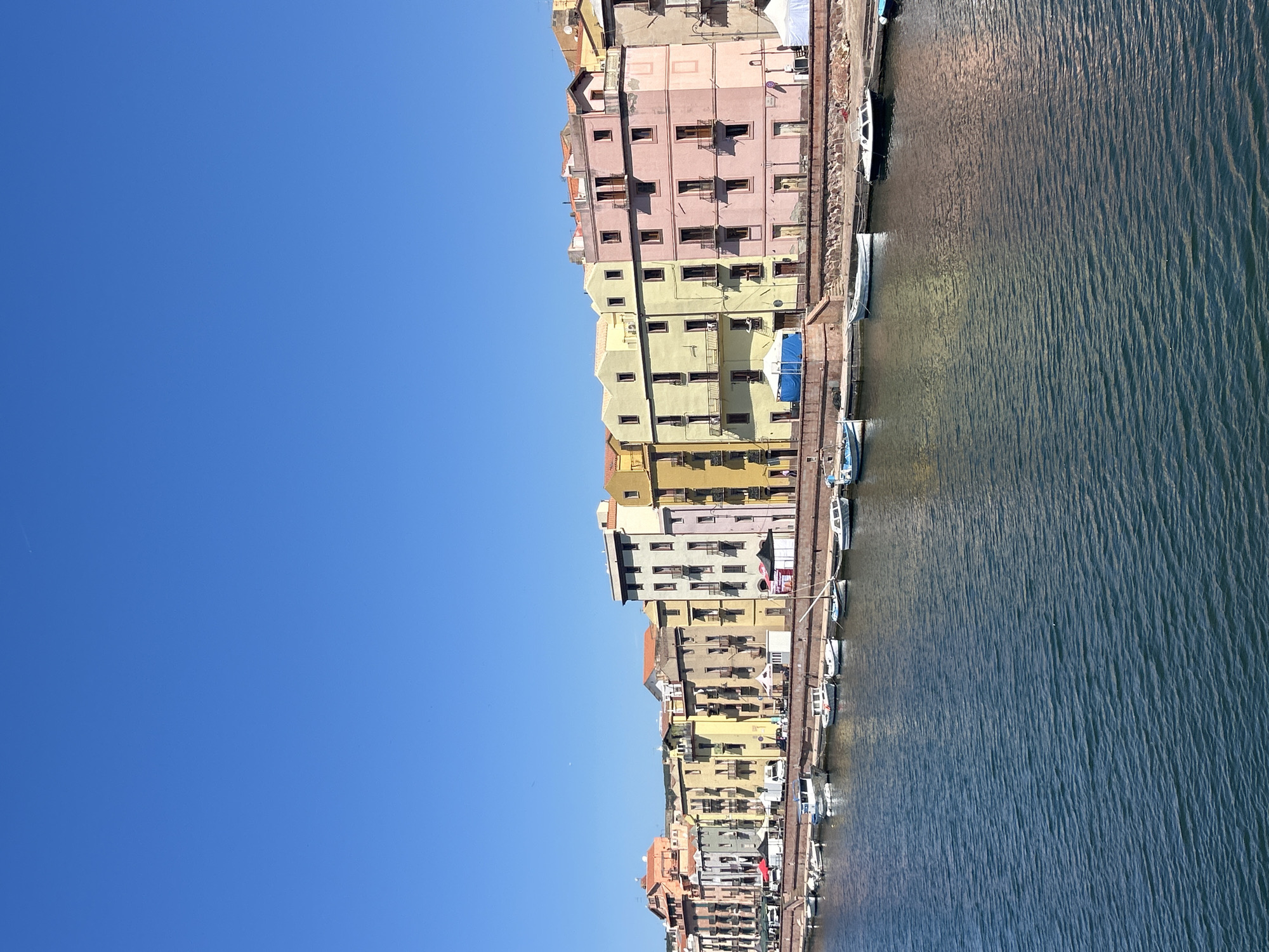 The image captures a row of colorful buildings along the Temo River in Bosa, Italy, under a clear blue sky. The buildings are various shades of yellow, pink, and beige, with multiple windows and floors. Small boats are docked along the riverbank next to the buildings. The water in the river is a dark blue color with gentle ripples.