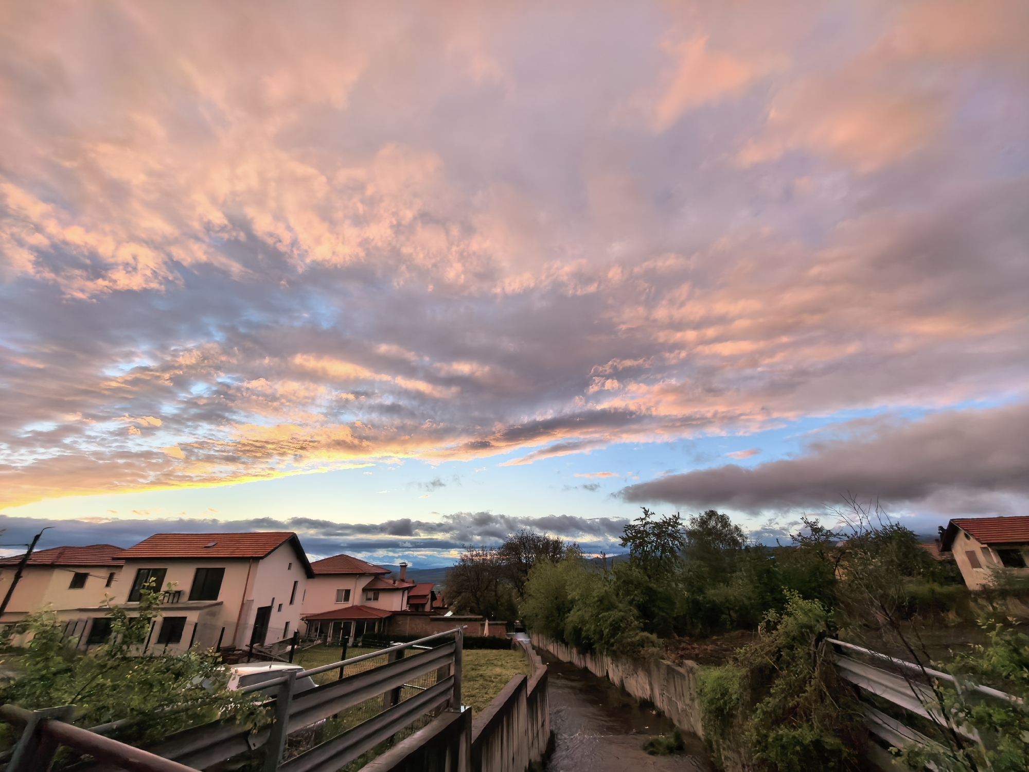 The image captures a vibrant sunset or sunrise scene over a residential area in Sapareva Banya, Bulgaria. The sky dominates the upper two-thirds of the frame, filled with dynamic clouds painted in striking hues of pink, orange, and light purple, interspersed with clear patches of blue sky. The warm, diffused light creates a dramatic and colorful overhead display.
In the midground, several houses with distinct red tile roofs and light-colored facades are visible, primarily on the left side of the frame. These houses appear modern, with some possibly still under construction or recently built, featuring dark-framed windows and balconies. Dense green vegetation, including various trees and bushes, lines a narrow concrete channel or canal that runs from the mid-right towards the foreground. Water is visible flowing within this channel. A metal guardrail extends across the lower left and center of the foreground, providing a visual barrier.
In the distant background, faint silhouettes of hills or mountains can be seen beneath the colorful horizon. There is no visible human activity or text in the image. The scene conveys a serene and picturesque moment, highlighting the beauty of the natural light over a quiet settlement.