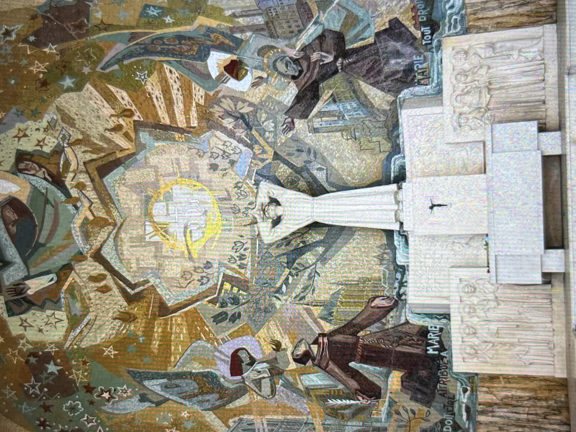 The image depicts an artistic tapestry or mural, possibly located in Tinaquillo, Venezuela, showcasing a religious theme. Central to the composition is a figure in a long white robe, possibly a depiction of the Virgin Mary. Two figures in brown monastic robes are positioned above and below her, seemingly reaching out. The background is a complex arrangement of geometric shapes, stars, and stylized buildings, creating an abstract cityscape. At the right edge, a sculpted stone element with text in French "MARIE" and "Tout Droit", and a smaller Latin Cross are present. The overall environment has a textured, tapestry-like appearance, blending religious imagery with architectural and abstract elements.