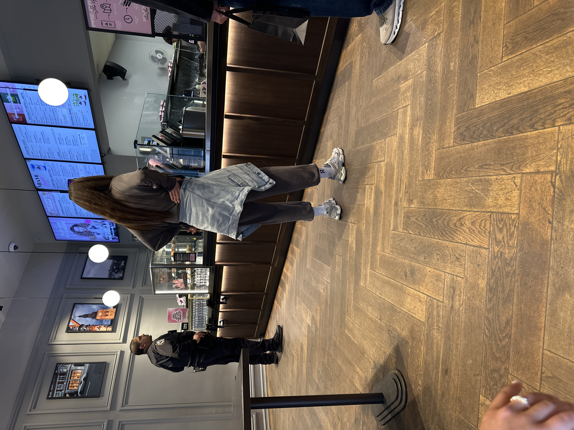 The image depicts an indoor scene within a modern cafe or juice bar, likely located in New York, United States. The establishment features light brown herringbone wood flooring and light-colored walls adorned with framed pictures and circular white light fixtures. A long, dark wooden counter, complete with display cases and a service area, runs along one side of the room.
Several people are present in the space. In the foreground, a person with long brown hair, wearing a gray top, dark pants, and a light denim jacket, stands facing the counter, appearing to wait in line. They are wearing white athletic shoes with white socks that display the letters "bc." To the left, a person in a dark uniform, resembling a security guard or staff member, stands near a table. Partially visible in the upper right is another customer's leg and white athletic shoe, while a hand with a ring, possibly belonging to the photographer, is visible in the lower right. A staff member can be seen working behind the counter.
The primary activity is people waiting at the counter, indicative of a typical cafe or quick-service setting where customers order or pick up food and beverages.
Notable details include a small white fan positioned behind the counter and a black stanchion with a rope barrier in the foreground. Menu boards prominently display various offerings, including "SPECIALTY COFFEE - ONYX," "SIGNATURE JUICES," "FUNCTIONAL SMOOTHIES," "SEASONAL DRINKS," and "PREMIUM SANDWICHES," with calorie information listed next to many items. A sign above the counter advertises "PRE-ORDER" options and contains text like "Love You," likely part of the brand's name. A smaller sign on the counter mentions "TIPS" and details a potential payment policy related to sales percentage. The interior lighting suggests it is daytime.