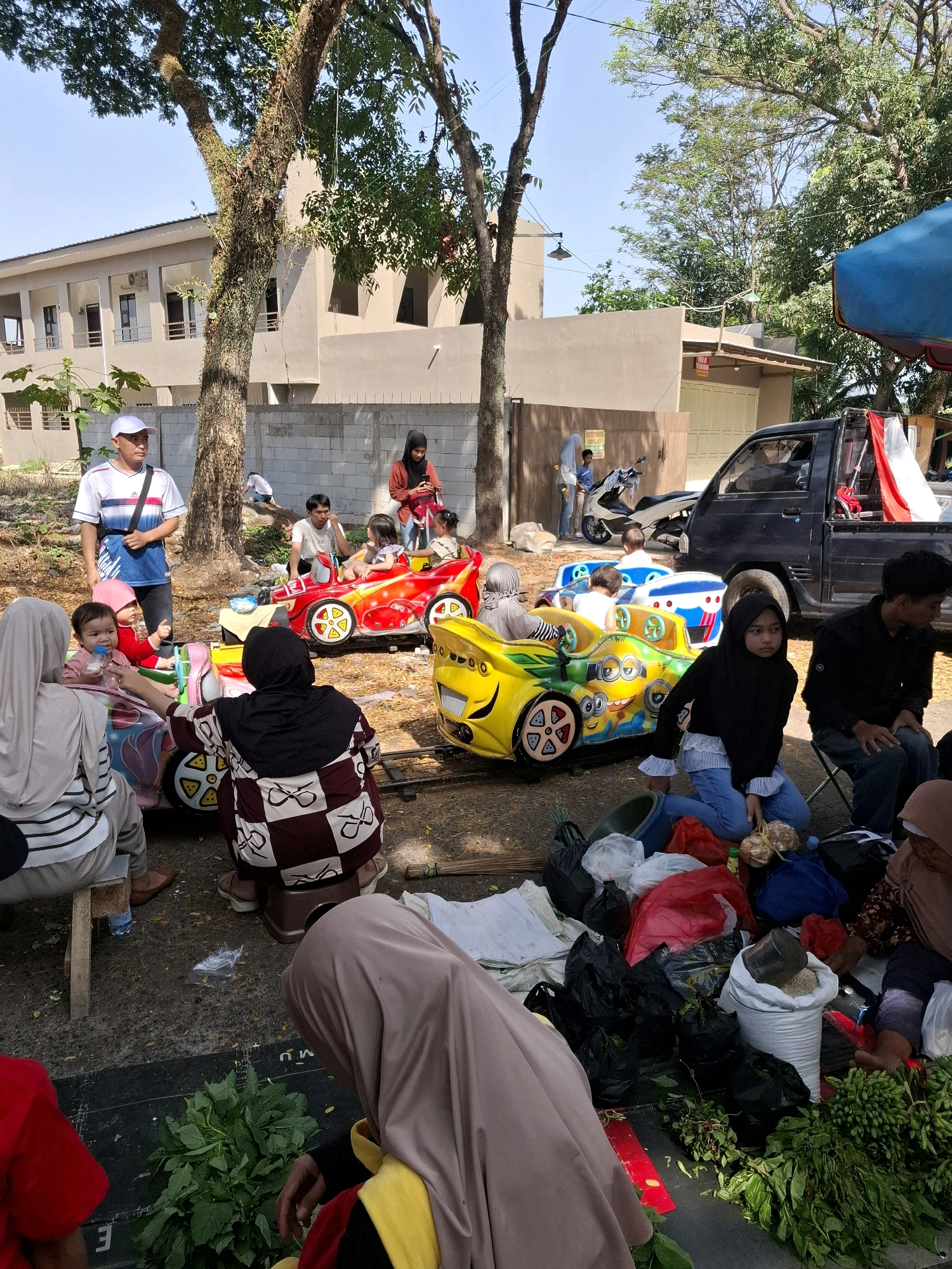 This image depicts an outdoor scene in Karangtengah, Indonesia, likely a local market or recreational area. In the foreground, several people, including women and children, are gathered around small amusement park rides shaped like cars. One child is being fed from a bottle. The rides are colorful, with one designed like a red sports car and another featuring cartoon characters.
In the background, a man in a white cap and t-shirt stands near a tree, observing the scene. Further back, a modern building with several windows and a fence can be seen. A black pickup truck is parked on the right side of the image, with an Indonesian flag prominently displayed in its bed. Various bags and bundles of produce, likely for sale, are spread out on the ground in the foreground and midground, suggesting a market environment. The ground is covered with dirt and scattered leaves. The overall atmosphere appears to be casual and family-oriented, with a mix of leisure activities and commercial transactions.