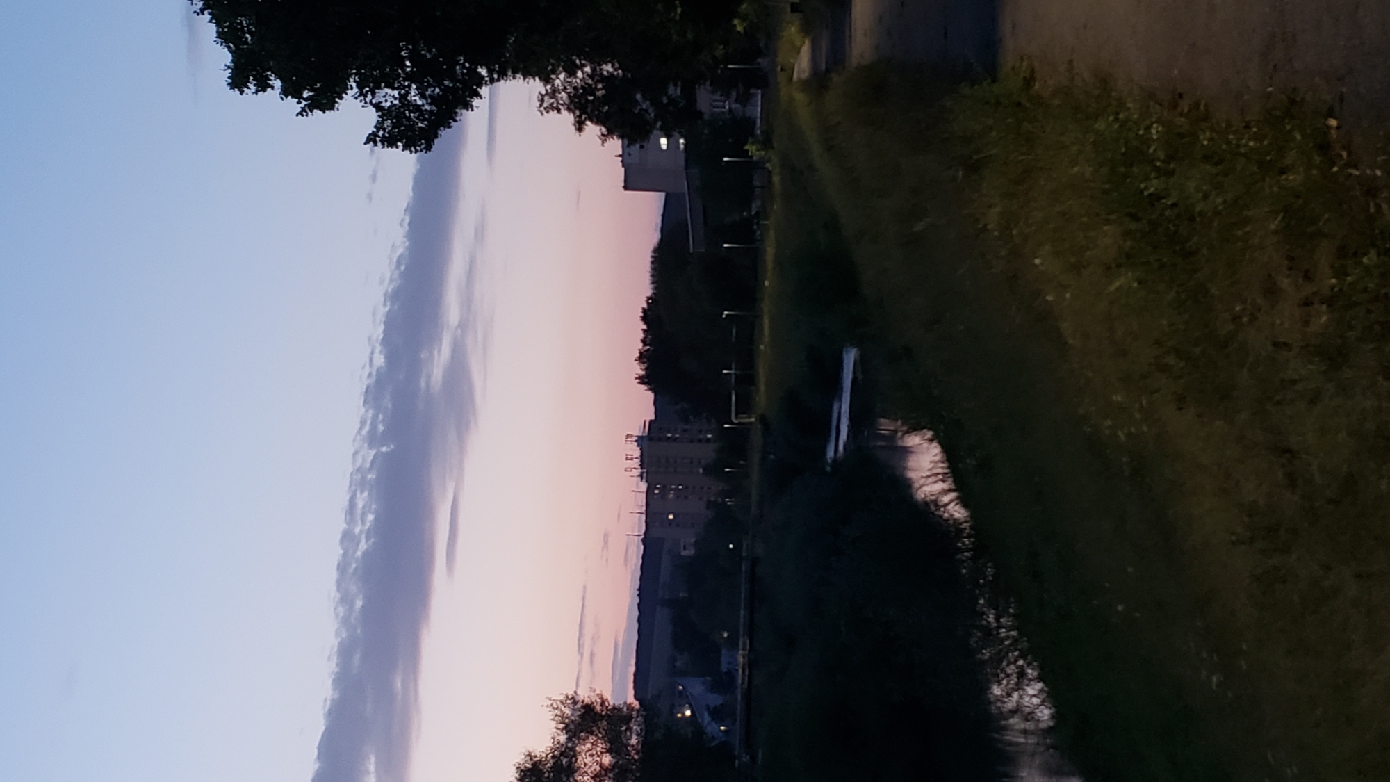 This image captures a twilight scene of a riverbank and residential buildings in Prešov, Slovakia. The sky is a soft gradient of pale blue and pink, with a few wispy clouds. A dark, tree-lined embankment runs along the left side of a calm river, reflecting the fading light. On the right bank, a paved path winds alongside a grassy slope. In the distance, several apartment buildings and other structures are visible, some with illuminated windows. Streetlights are spaced along the path and in the open grassy areas, indicating a public space. The overall atmosphere is peaceful and serene, suggesting early evening in a suburban or urban park setting.