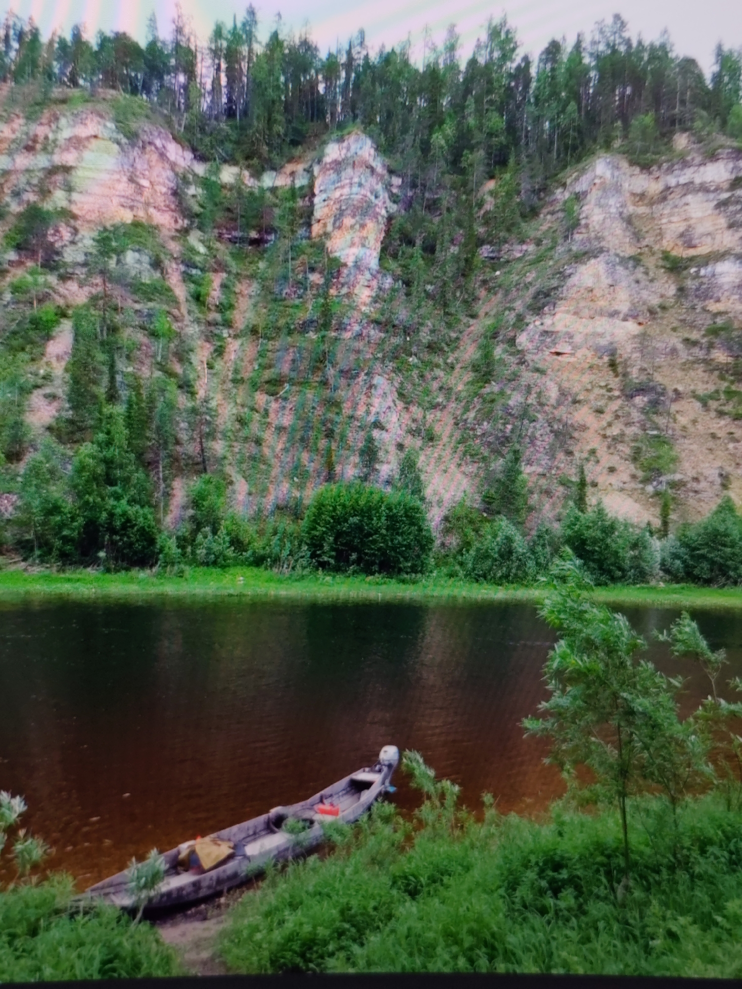 The image depicts a tranquil river scene bordered by a rocky cliff face covered in sparse vegetation and trees. In the foreground, a canoe rests on the grassy bank of the river, with a yellow tarp and a red object visible inside it. The water is dark and reflects the sky and surrounding greenery. The cliff face exhibits layers of rock with patches of green moss and trees growing from its crevices. The top of the cliff is lined with a dense forest. The scene appears to be a natural landscape, likely a wilderness area, and there is no indication of it being located within the city of Syktyvkar, Russia.