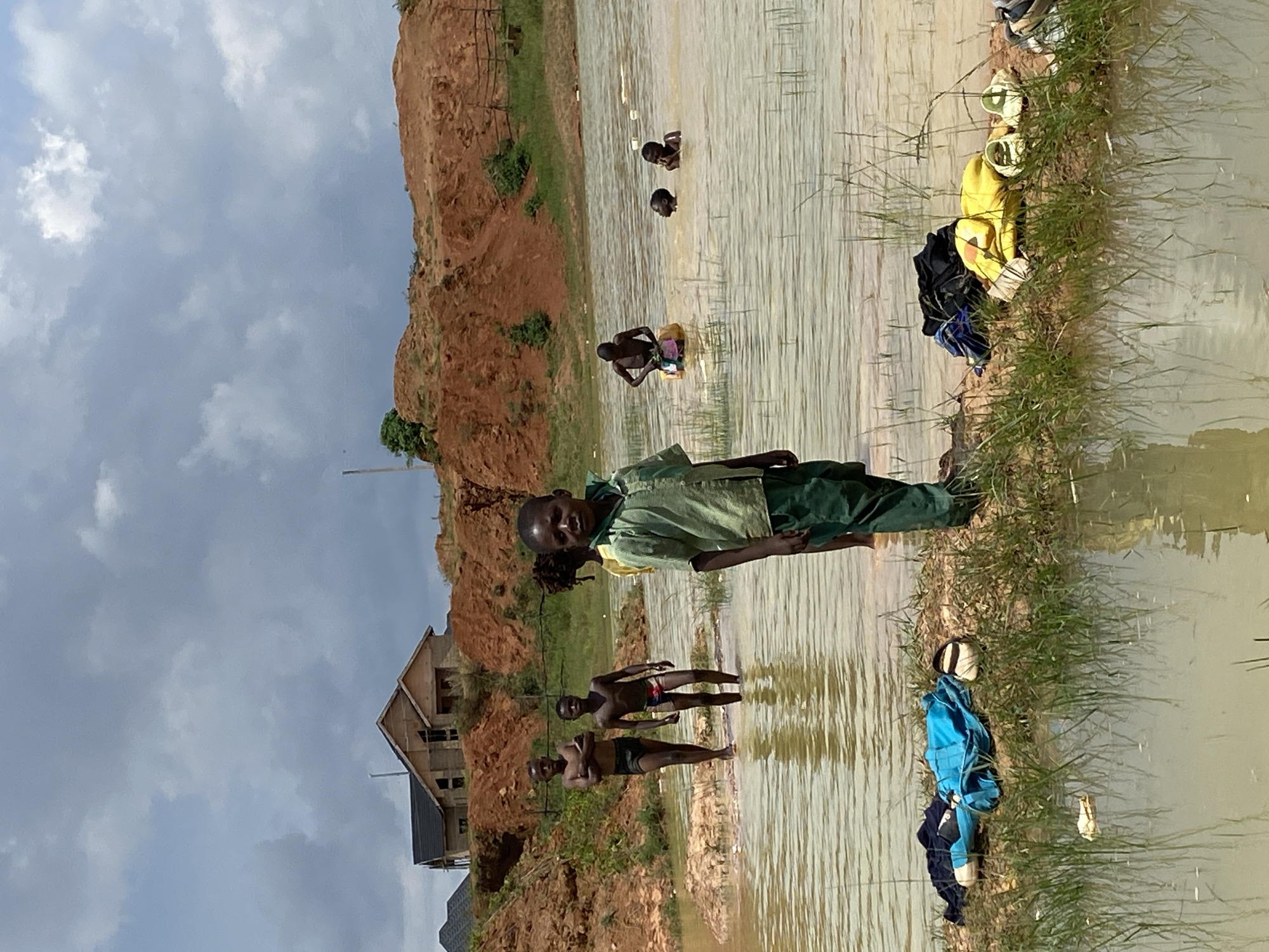 In Bar Jirgi Summa, Nigeria, on a cloudy day, several children are playing in a body of muddy water, possibly a large puddle or pond. In the foreground, a young boy in a green collared shirt and dark pants stands facing the camera, looking directly at it. Behind him, two other boys wade in the water, one with arms crossed and the other with a stride. Further back, a group of children are bathing and playing in the water. On the bank to the left, clothing and shoes are neatly placed on the ground. In the background, a construction site is visible with partially built houses and mounds of reddish-brown earth. The environment appears to be a developing area with natural and man-made elements.