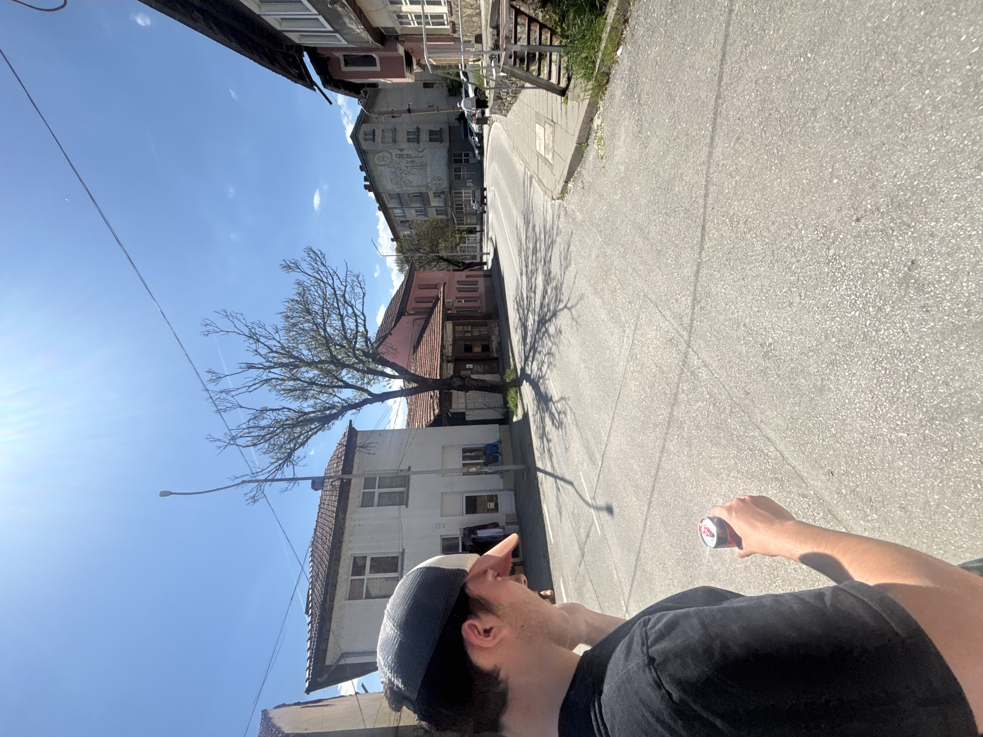 The image shows a person standing on a paved street in Troyan, Bulgaria, looking toward a line of buildings on a slight incline. The sky is blue with a few clouds. A bare tree stands on the left, casting a shadow on the street. The person is wearing a black T-shirt and a baseball cap and holding a can in their right hand. Buildings of varying ages and construction styles line the street, including some with tiled roofs and multiple stories.
