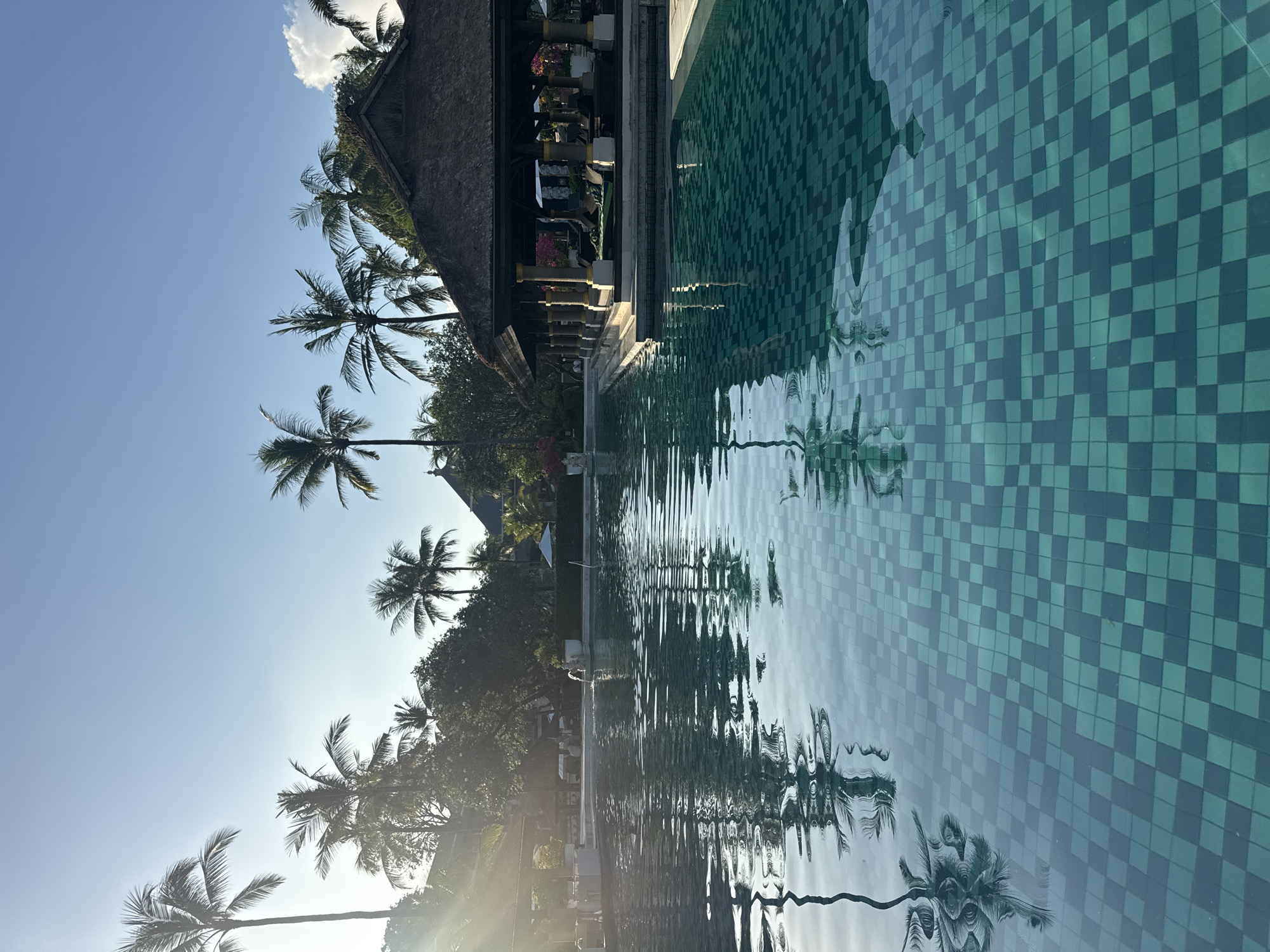 The image depicts a swimming pool in Kuta Selatan, Indonesia. The pool is tiled with small, square, green and blue tiles. The water is slightly rippled, reflecting the surrounding palm trees and a building with a thatched roof. The sky is clear blue with a few clouds. Sunlight is visible, casting a glare on the lower left of the image.
