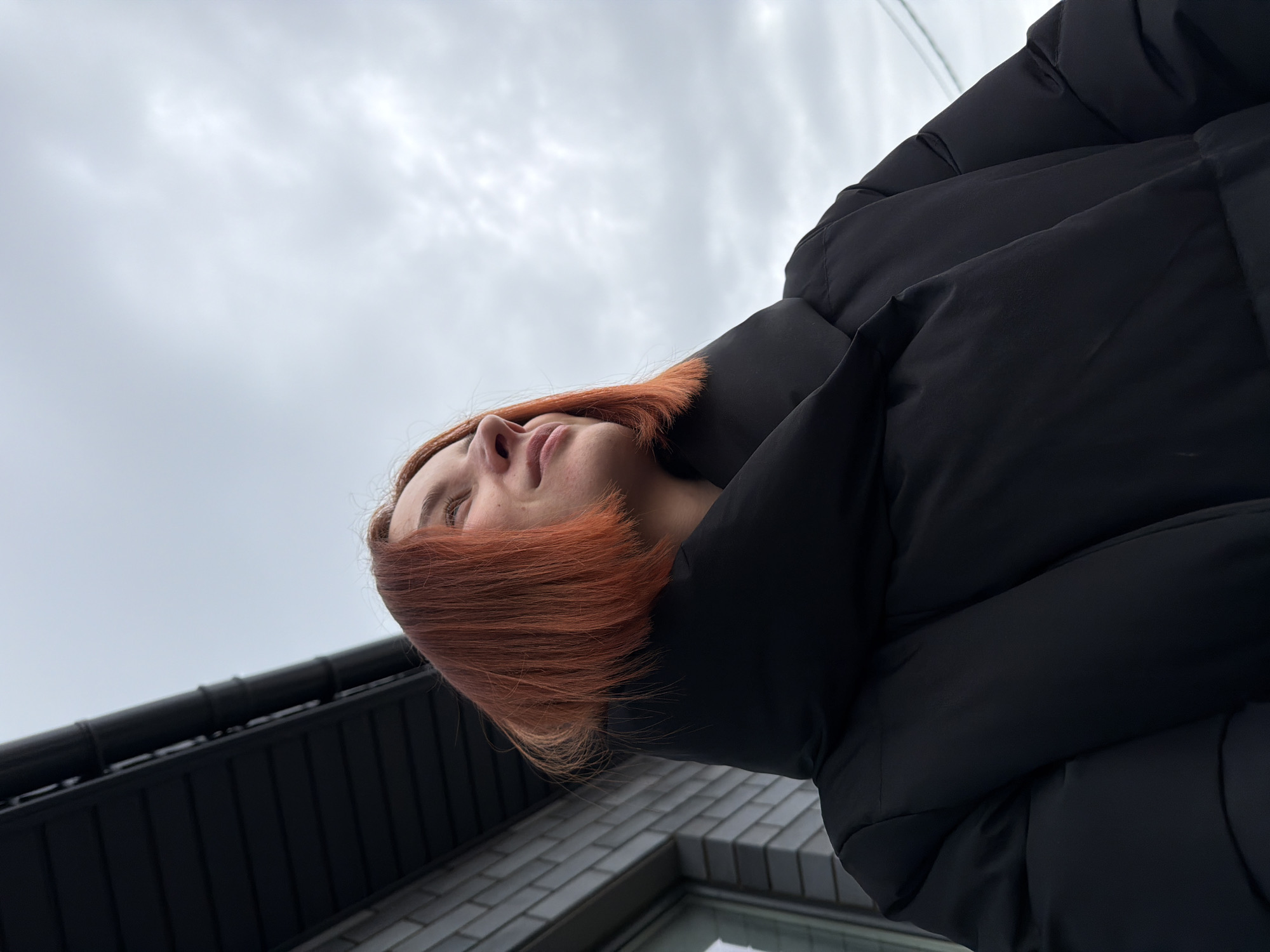 A low-angle shot of a person with short, vibrant orange-red hair, likely a woman, standing outside. She is wearing a large black puffer jacket. The background includes a grey, cloudy sky, a building with dark roofing and light-colored brick walls.