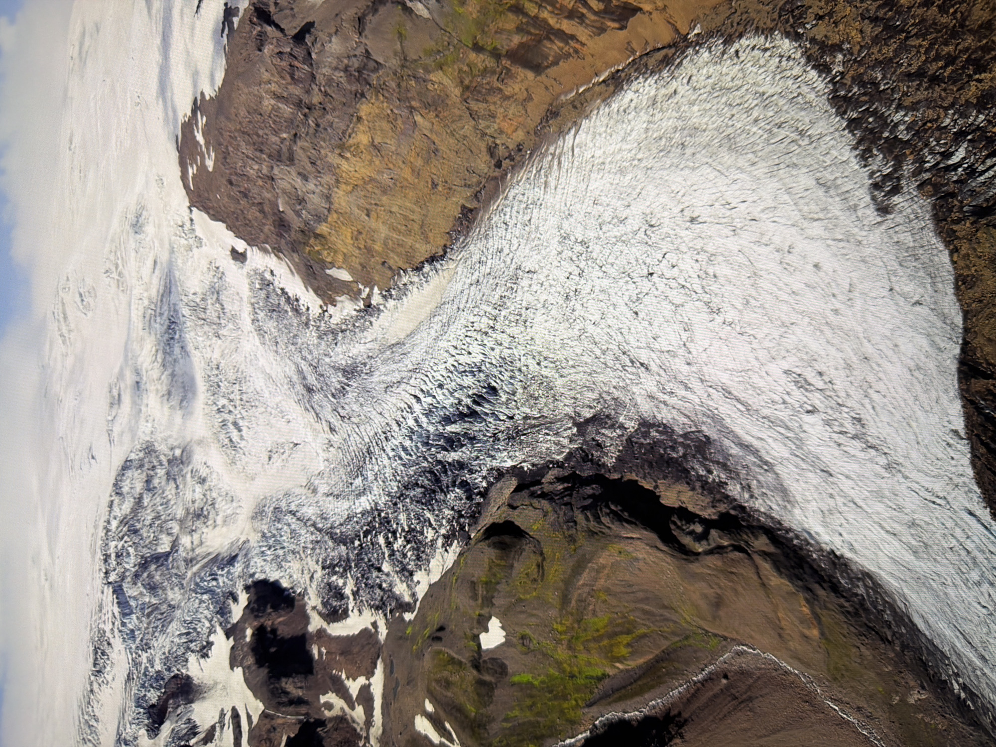The image is an aerial view of a vast glacial landscape, likely in Iceland or a similar arctic region. A large, textured glacier dominates the scene, its surface marked by intricate patterns of crevasses and ridges, indicating slow movement and ice deformation. The glacier flows down from a higher elevation, where it appears to merge with a more extensive ice field. Surrounding the glacier are steep, rocky mountainsides and valleys. Some of these areas are covered in snow and ice, while others reveal exposed earth and rock, with patches of green vegetation suggesting the presence of moss or low-lying plants in more sheltered areas. The color palette consists primarily of whites and blues from the ice, contrasted with the earthy browns, grays, and occasional ochre and green of the surrounding terrain. The lighting suggests it is daytime, with shadows casting definition on the glacial surface and mountainous features. There are no visible signs of human structures or activity. The image does not depict the city of Kyiv, Ukraine.