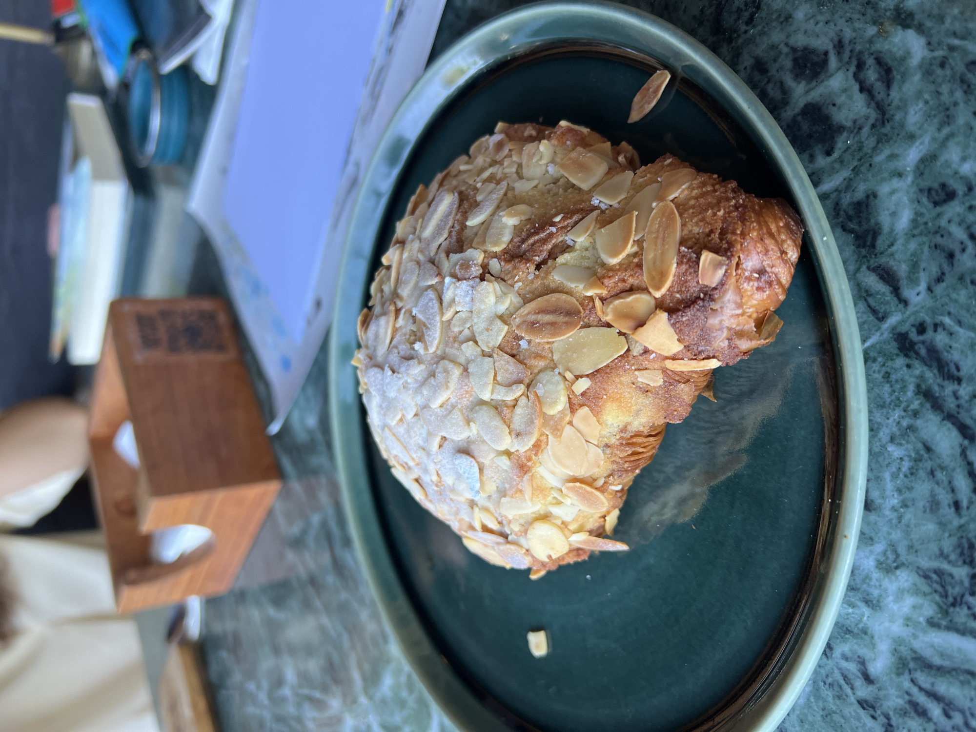 The image shows an almond croissant on a dark green plate, placed on a surface that appears to be marble or a similar material with a mottled pattern. A small, rectangular wooden object is visible on the left side of the frame, slightly out of focus. The background is somewhat blurred, but suggests other items on the table, including papers and containers. The overall scene suggests a cafe or restaurant setting. The location is Sukawati, Indonesia, but there are no elements in the image that definitively confirm this location without prior knowledge.