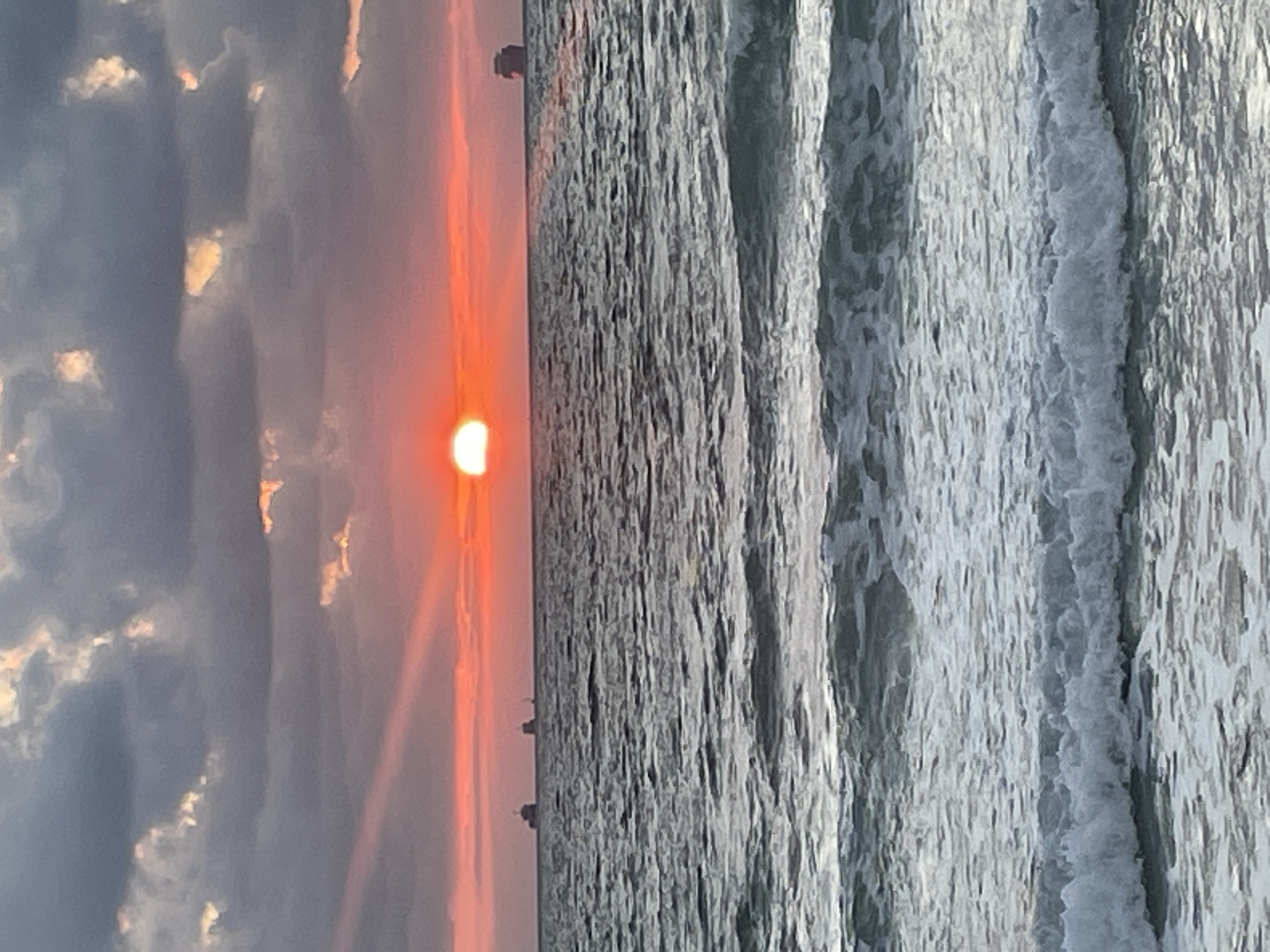 The image depicts a seascape at sunset, likely taken from a high vantage point, possibly a balcony or cliff overlooking the ocean. The sun, a vibrant orange orb, is partially obscured by clouds and is setting over the horizon, casting a warm glow across the sky. The clouds are a mix of dark grays and lighter, illuminated patches, suggesting a dynamic sunset with varying light conditions.
The ocean is a prominent feature, with a series of distinct waves crashing against what appears to be a rocky or man-made shoreline. The texture of the water is rough and dynamic, with white foam cresting the waves. In the distance, along the horizon line, there are faint silhouettes of what could be ships or structures, adding a sense of depth and scale to the scene.
The overall mood is serene and contemplative, evoked by the dramatic interplay of light and shadow during sunset. The colors are rich and warm, with the orange of the sun contrasting with the cool grays and blues of the ocean and sky. The image seems to capture a moment of natural beauty and tranquility. The location context provided suggests this scene is from Haifa, Israel, a city known for its coastal beauty. No visible text is present in the image.
