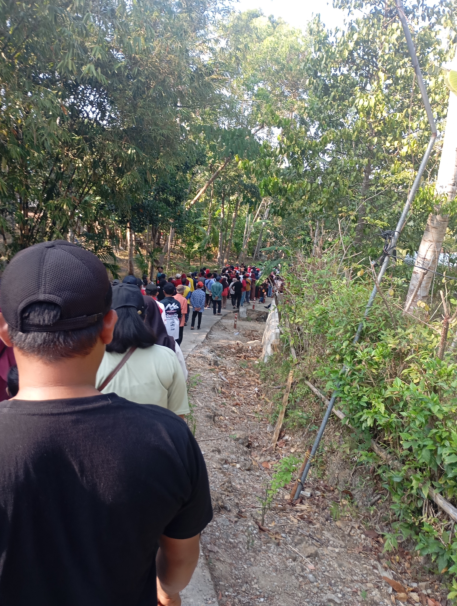 In Ketepung, Indonesia, a group of people are walking in a single file line along a path bordered by dense foliage and trees. The path appears to be made of gravel and dirt, with some paved sections visible. The individuals are dressed in casual clothing, with some wearing hats and t-shirts. The overall scene suggests an outdoor activity, possibly a pilgrimage or a guided tour, taking place in a natural, somewhat rural setting within the city. The lighting indicates it is daytime, with sunlight filtering through the leaves.