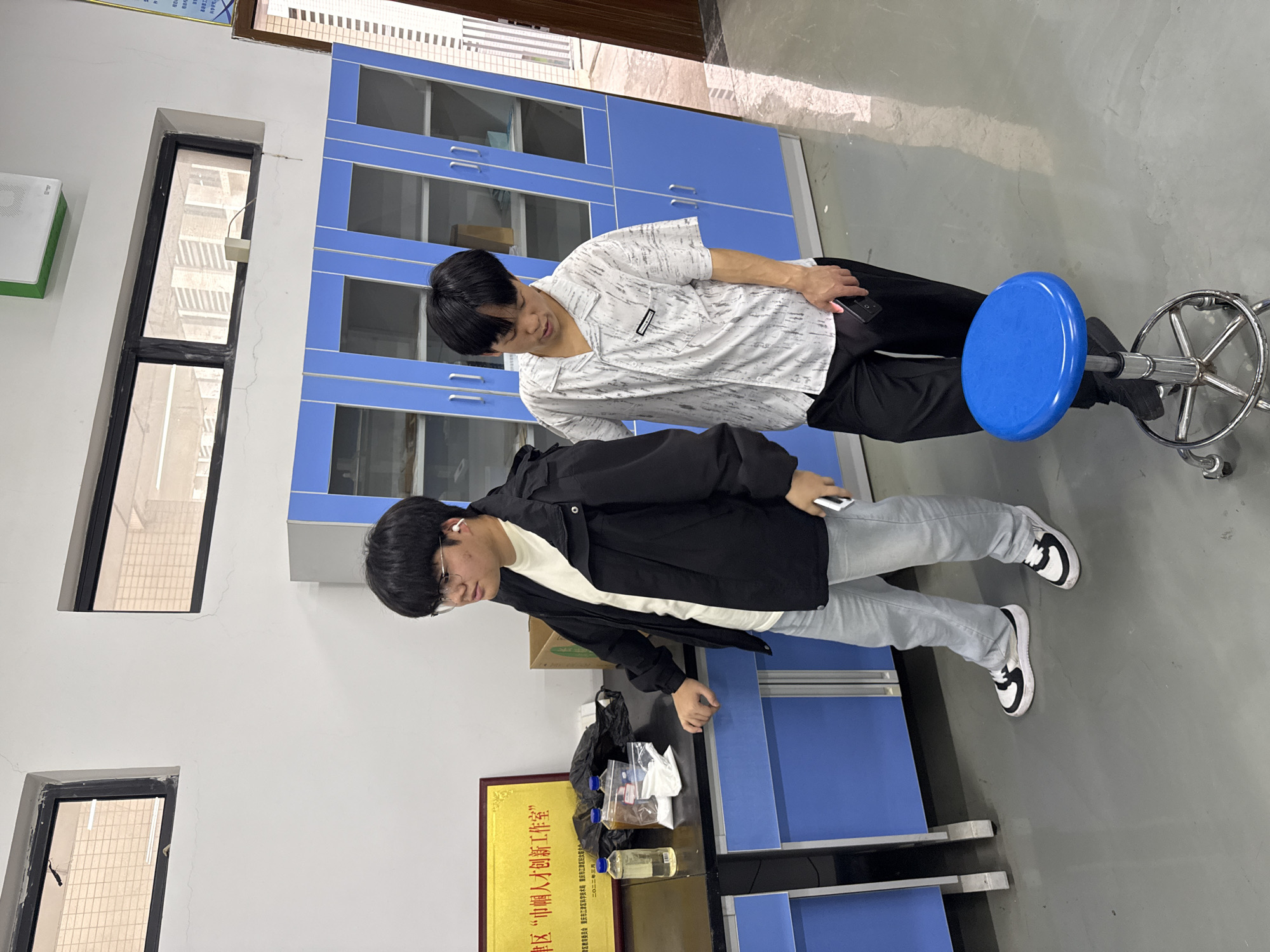 Two young men are in what appears to be a laboratory setting. The environment contains blue cabinets, laboratory equipment, and a stool. One man is standing, partially seated on the blue stool. He is wearing a patterned white shirt and black pants. The other man is also standing, wearing a white shirt, a black jacket, light blue jeans, and black and white sneakers. The lab environment suggests this might be a science or research facility. There is a plaque visible behind the man with the jacket, indicating that the lab might be located in the Jiangjin district of Chongqing, China.