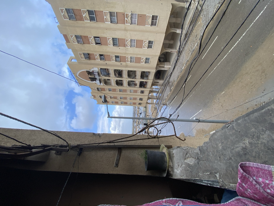 The image provides an elevated, oblique view of a street and urban landscape in Khan Yunis, Palestinian Territory. In the immediate foreground, to the left, a concrete ledge supports a black potted plant containing sparse green foliage, resting on a yellow saucer. A pink and white striped textile partially obstructs the far-left edge. Multiple black utility cables, some irregularly strung and attached to a building structure, crisscross the left side and upper portion of the frame.
The midground features an asphalt road, visibly wet with standing water and mud along its edges, featuring two dashed white lane markers. A grey lamppost with a light fixture stands on the right side of the road, extending upwards. Across the street, a multi-story residential building, light beige with patterned brown architectural details around its windows and balconies, dominates the right background. The ground floor of this building is elevated on a series of arched columns. Several balconies on the building appear to have exposed interiors or coverings, particularly in the central vertical section.
In the far background, beyond the street and the residential building, a large expanse of numerous temporary shelters, primarily white and grey, is visible, suggesting an encampment or informal settlement on what appears to be muddy ground. The sky is partially cloudy with patches of blue. No individuals or active interactions are discernible.