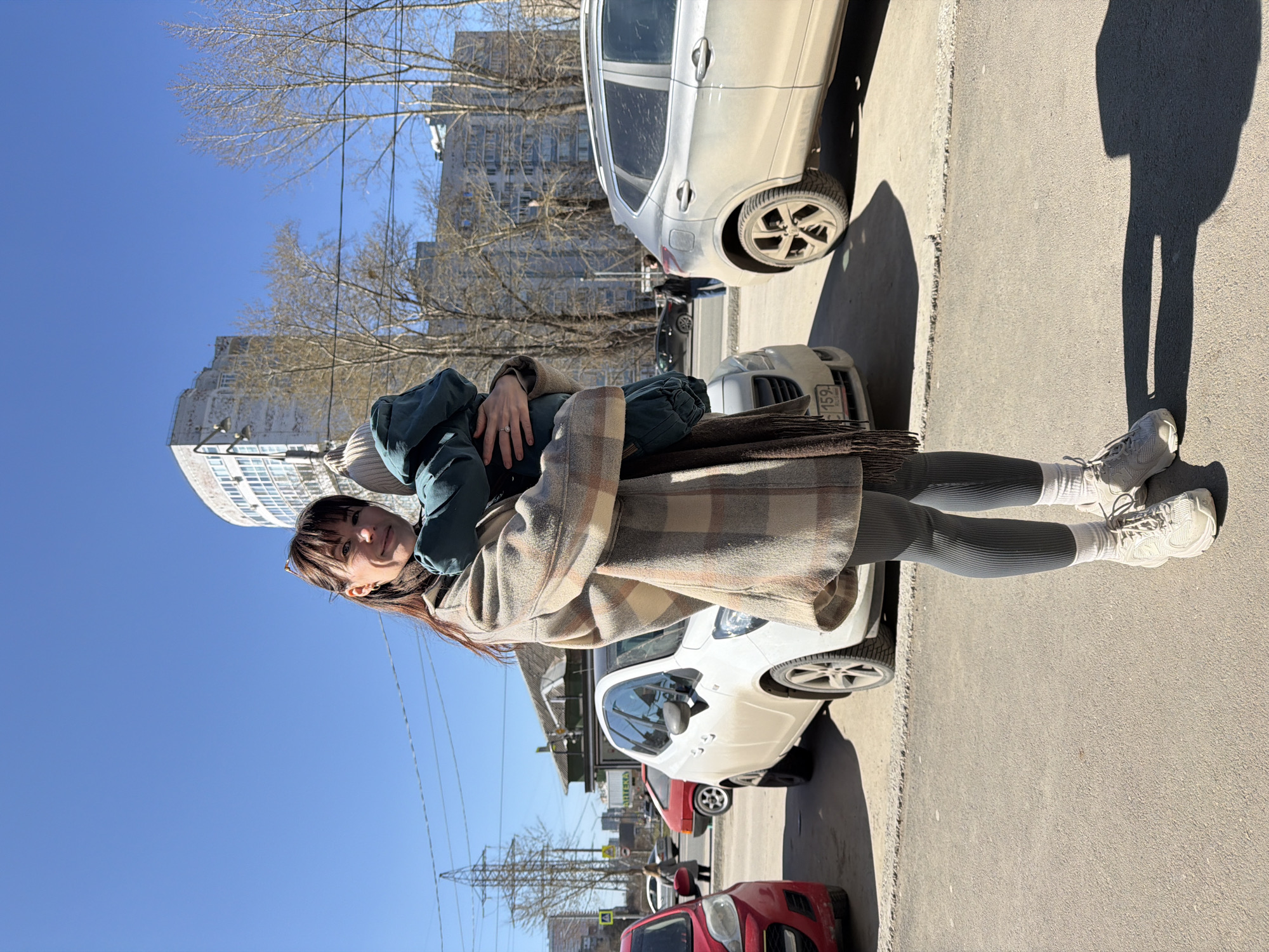 The image shows a woman standing on a sidewalk in Perm, Russia. She is wearing a plaid coat, gray leggings, white socks, and white sneakers. She is holding a child, who is wearing a dark green jacket and pants. The background includes parked cars, buildings, and trees. The sky is clear and blue.