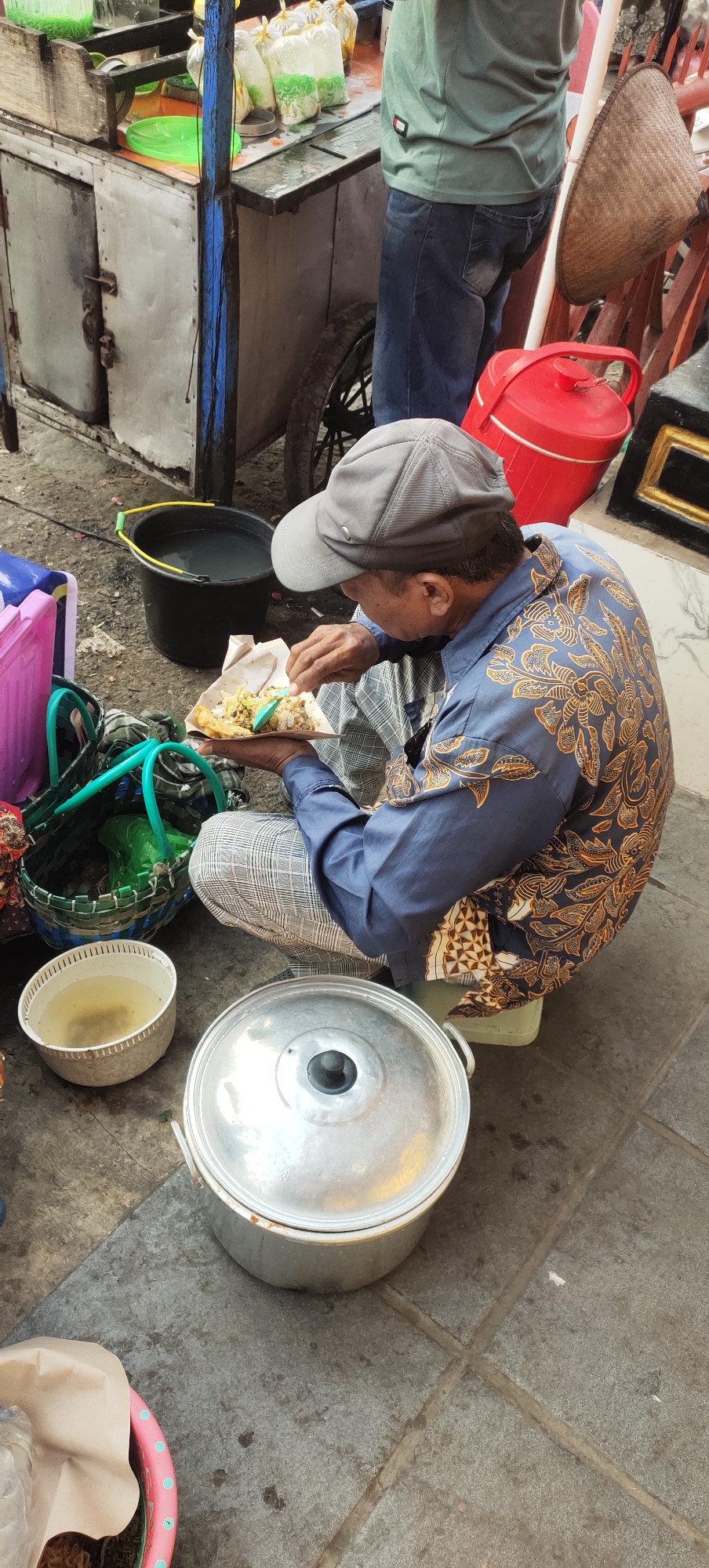 A man wearing a grey baseball cap and a dark blue patterned shirt is sitting on the ground, eating from a paper plate. He is also wearing plaid pants. In front of him is a large metal pot with a lid, and to his left is a white bowl containing liquid. Behind him is a food cart with various items, including bags of food and a large black bucket of water. To the right of the food cart, a red container and a woven hat are visible. The scene appears to be set on a paved area with some debris on the ground.