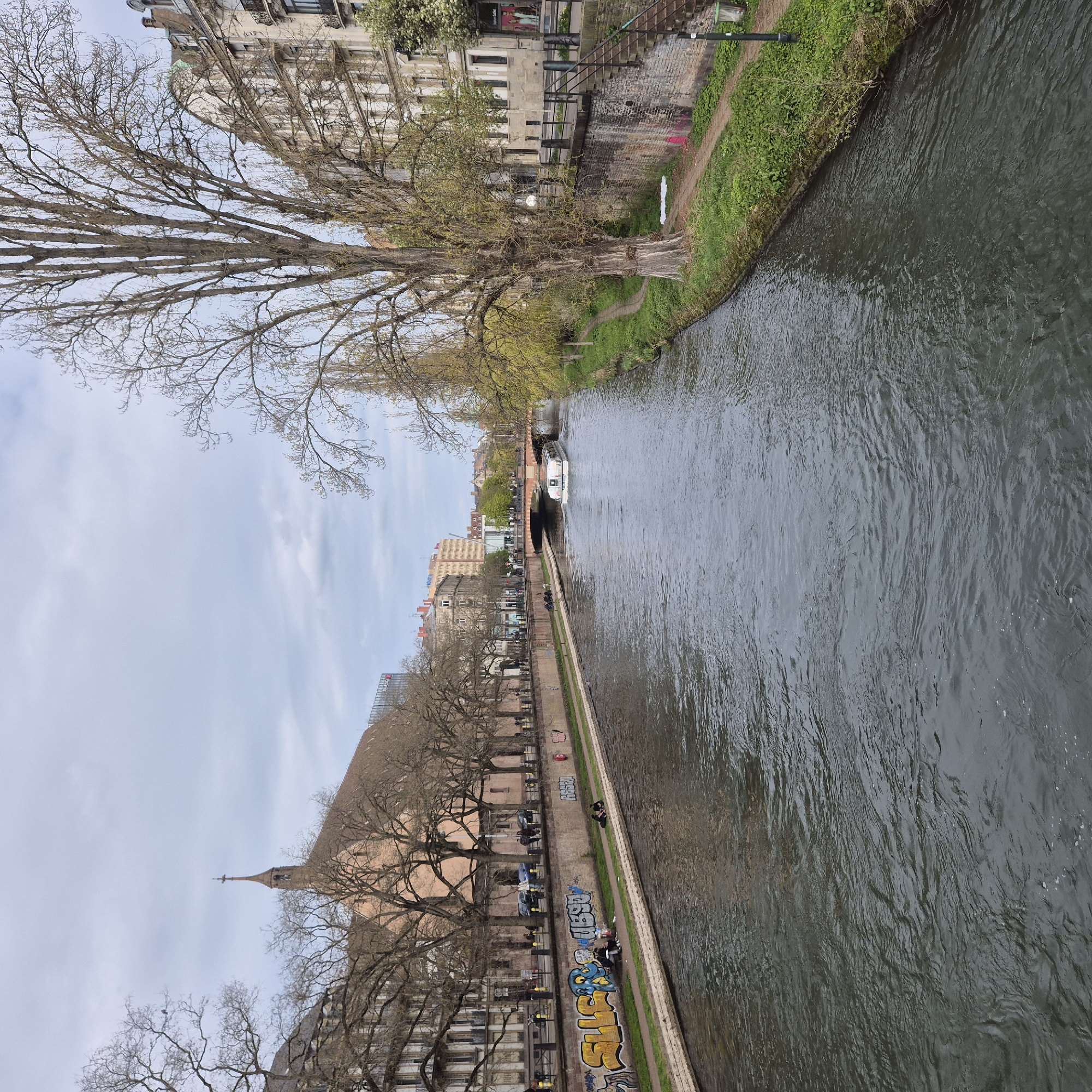 The image shows a canal flowing through Strasbourg, France. The canal's water is dark and rippled. A tour boat is seen traveling through a tunnel. On the left side of the canal, there is a narrow path lined with trees, a building, and a church with a tall steeple. People are seated along the path near graffiti. The sky is overcast.