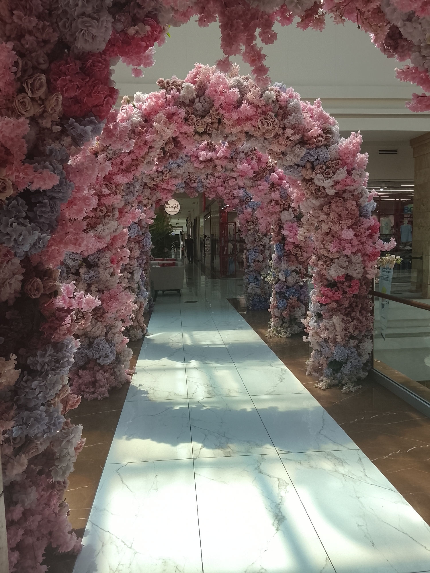 The image presents a perspective looking down a corridor adorned with a lavish floral archway. The archway is densely covered with artificial flowers, predominantly in various shades of pink, ranging from pale blush to vibrant rose, interspersed with clusters of light blue or lavender flowers. These appear to be a mix of rose-like blooms and smaller, clustered flowers resembling cherry blossoms or hydrangeas, creating a lush, immersive tunnel effect.
The pathway beneath the archway features a central section of bright white marble tiles, characterized by subtle grey veining and a highly polished, reflective surface. This white pathway is flanked on both sides by a darker, glossy brown or bronze-colored marble or stone flooring. Strong overhead lighting casts distinct, elongated reflections and shadows onto the white marble, indicating a well-lit indoor environment.
Beyond the floral archway, the corridor extends into a larger indoor space, likely a shopping mall or public venue. Storefronts or display areas are visible in the background, with a white sofa or bench partially discernible on the left side further down. A faint, dark figure, possibly a person, can be seen walking in the distance on the right.
A circular white sign, partially obscured by flowers, hangs within the archway on the left. It reads "In tempo" with smaller text below, possibly "Time to Enjoy". Further down the corridor on the right, some blurry text, including Arabic script and numerals like "2000," is visible on a display. The overall ambiance is one of beauty and elegance, suggesting a decorative installation for a special occasion or to enhance the aesthetic of the venue during daytime.