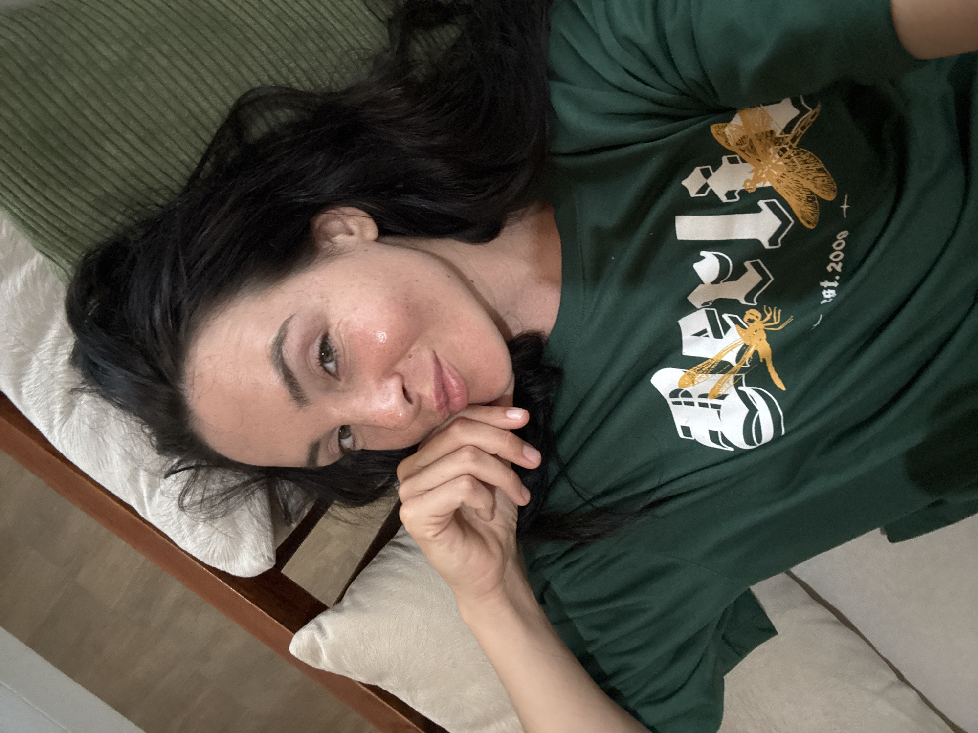 A high-angle, medium shot shows a woman with dark hair lying on her side on a couch. She is wearing a green t-shirt with a graphic design featuring dragonflies. Her left hand is resting on her chin and her right arm is bent at the elbow. The woman is looking towards the camera with a slight smile. The couch appears to be in a living room setting, with pillows visible behind her. The floor in the background is visible and appears to be made of wood. The scene does not provide any specific clues to pinpoint the location to North Kuta, Indonesia.