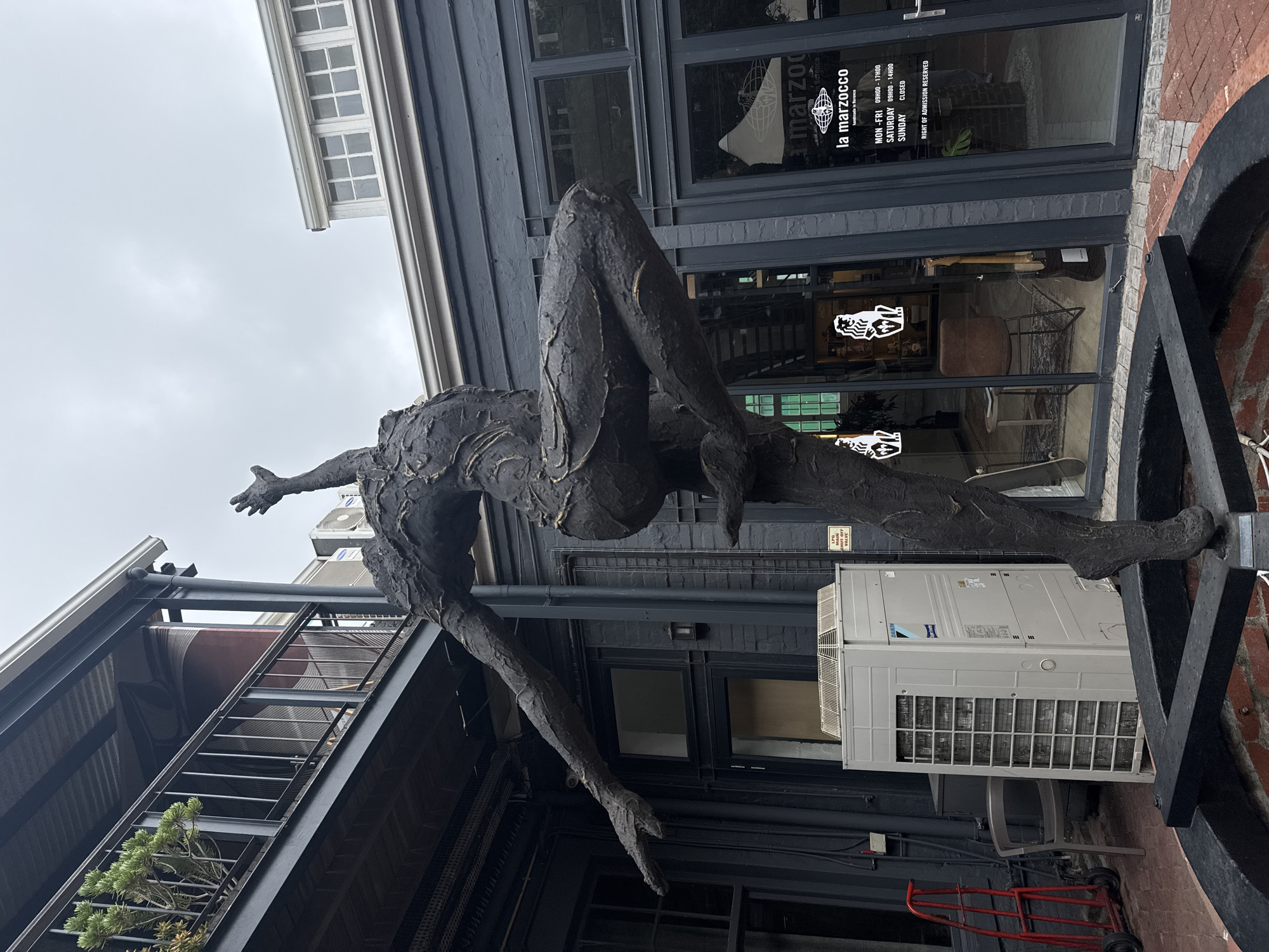 The image depicts a bronze sculpture of a human figure in an acrobatic pose. The sculpture is located outside a building with dark gray walls and windows. The building has a sign that reads "la marzocco", indicating a coffee shop, with the text “handmade in Florence” underneath. There is an air conditioning unit on the wall, along with a metal platform for the sculpture to stand on. The sky is overcast. The coffee shop is in the city of Cape Town, South Africa.