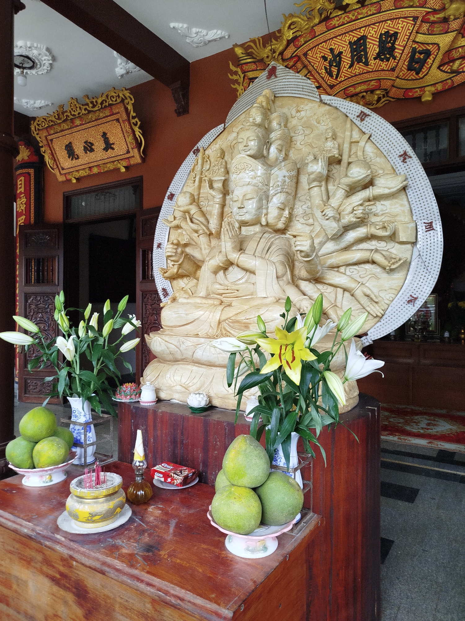 A medium shot of a Buddhist shrine in Pleiku Ward, Vietnam, displays an ornate wooden table holding various religious offerings. On the left, a white bowl supports several green pomelos, with a vase of white lilies behind it. Next to this is a yellow incense holder with several red sticks, a small brown bottle, and a package of red cakes. On the right, another white bowl also containing pomelos is next to a vase of white and yellow lilies. Behind the table is an intricately carved Buddha statue with multiple heads and arms, set against a white halo. Above the statue, there are two decorative plaques with gold frames and Asian characters. The background shows a partly open wooden door.