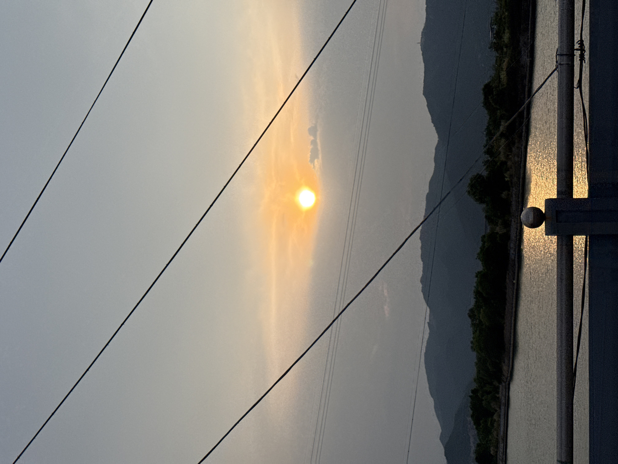 The image captures a serene natural landscape at either sunrise or sunset, viewed from a slightly elevated vantage point. The sky, occupying the upper portion of the frame, transitions from a pale, warm golden hue around the central, luminous sun to a soft, cool greyish-blue towards the edges. The sun, a bright, distinct orb, casts a soft glow, suggesting a hazy atmosphere or thin clouds.
Several dark, thin power lines diagonally traverse the sky, stretching from the upper-left to the lower-right, adding a man-made linear element to the vast expanse.
In the lower half of the image, a calm body of water—likely a river or a lake—reflects the warm light of the sun, creating a shimmering, golden path across its surface. Beyond the water, a dense band of dark green trees or foliage lines the distant bank. A range of majestic mountains, silhouetted in dark blue-grey tones, rises behind the trees, their peaks fading into the atmospheric haze of the horizon.
On the far right, a dark, metallic structure, possibly a part of a railing or a lamppost, stands prominently, partially obscuring the view of the water. A distinctive spherical finial adorns this structure, catching the reflected light from the water. The scene evokes a peaceful and expansive natural setting, characteristic of a heritage area like Nam Natural Heritage Area Trang in Vietnam. No text is visible in the image.