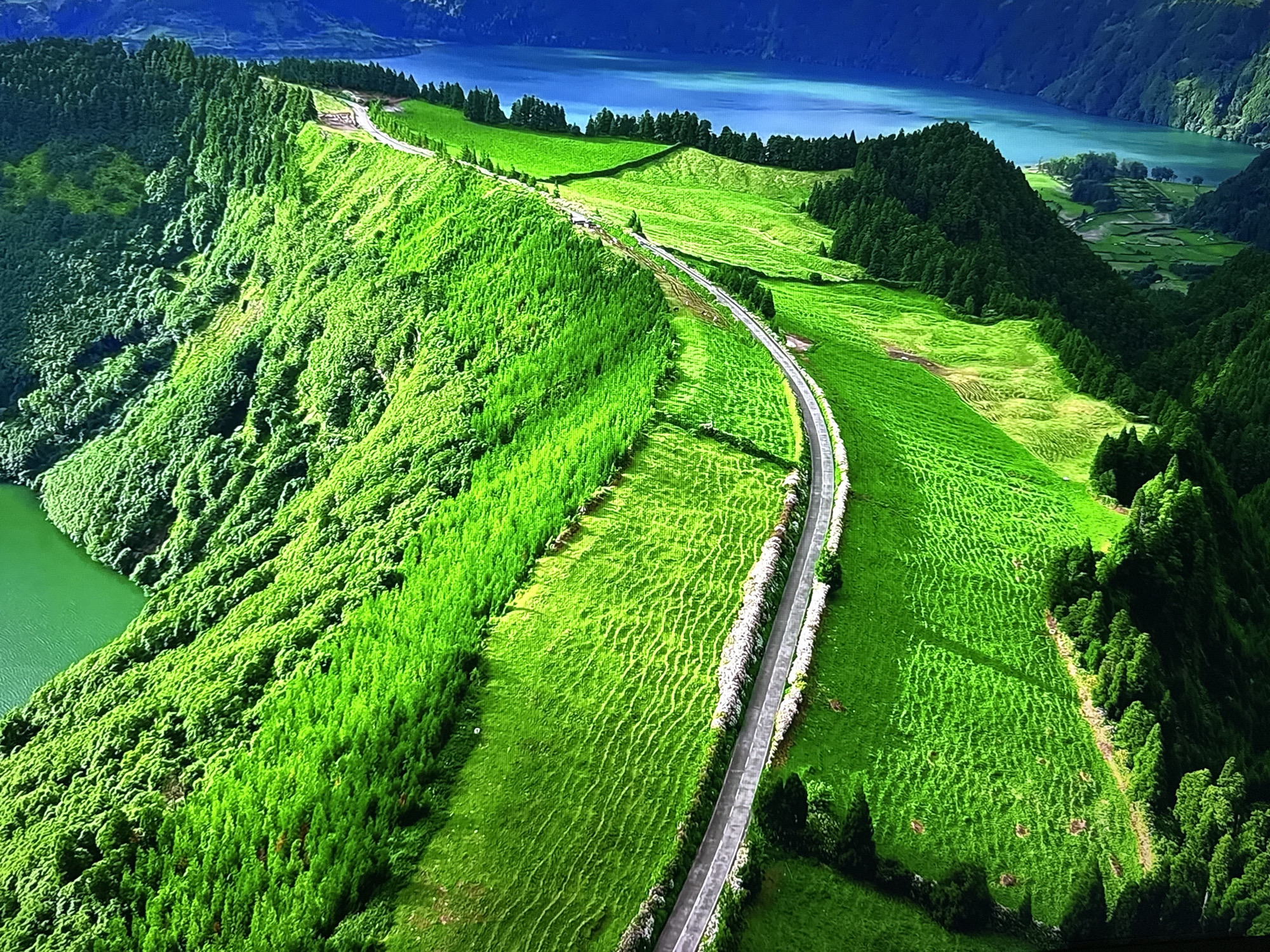 An aerial, high-angle, long shot captures a verdant, mountainous landscape dotted with lush green trees and vibrant green fields. A winding asphalt road, bordered by stone walls, meanders through the terrain. In the background, a tranquil, blue body of water reflects the sky, nestled between rolling hills. The overall scene is one of natural beauty and serenity. This image is not of Burgas, Bulgaria, but rather depicts a natural landscape, likely a volcanic crater lake, given the topography and vegetation.