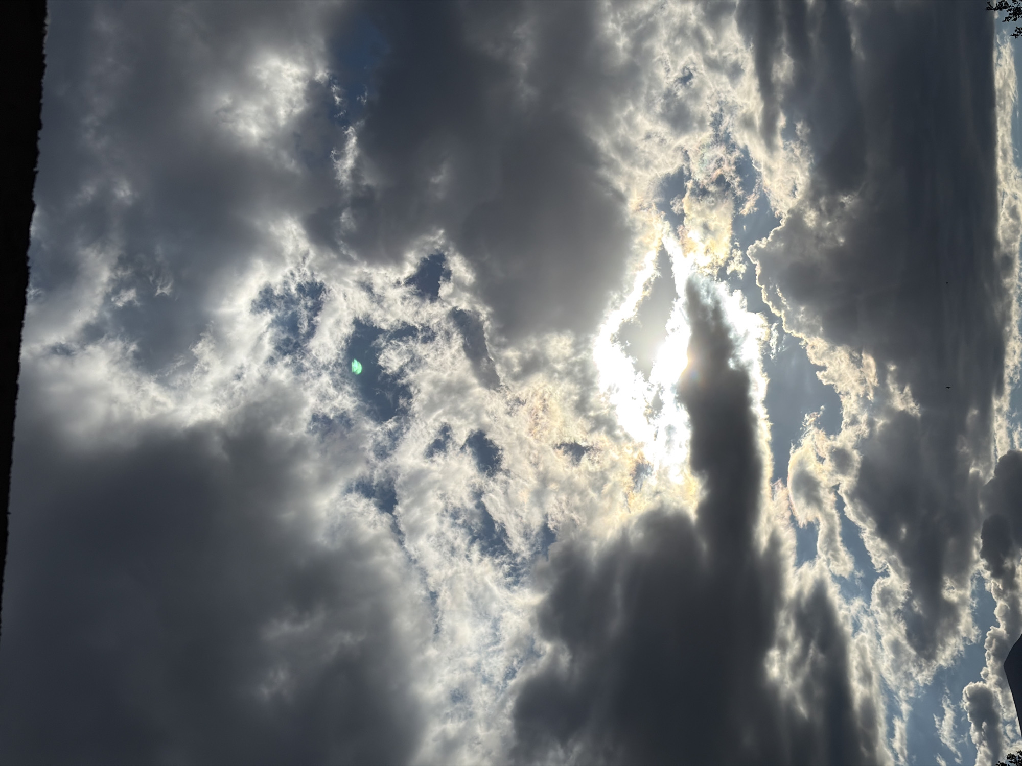 The image shows a dramatic sky with a mix of dark, heavy clouds and bright, sunlit areas. The sun is partially obscured by clouds, creating a powerful contrast of light and shadow. In some parts of the sky, the sunlight appears to refract through the clouds, creating iridescent or rainbow-like effects. The overall impression is one of a dynamic and possibly stormy sky, or perhaps the aftermath of a storm. The bottom edge of the image shows a dark, silhouetted line, likely indicating the presence of a mountain range or a dark structure against the sky. There are no discernible people or specific objects in the sky that would indicate a particular event or activity. The image is likely a landscape photograph focused on atmospheric conditions.