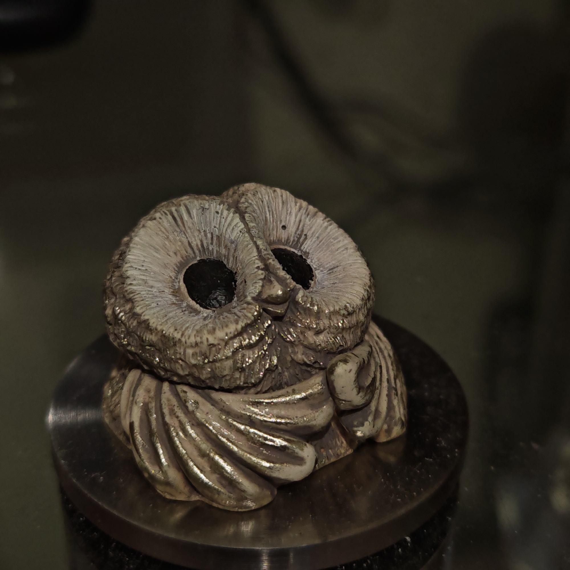 A close-up shot depicts a small, carved, owl figurine with large, dark eye sockets. The figurine appears to be made of a light-colored material, possibly wood or clay, with detailed texture suggesting feathers. The figurine rests on a circular, metallic base. The background is dark and blurry, suggesting a dimly lit indoor setting. There is no indication that this image was taken in the city of Novo-Nikol'skoe, Russia.