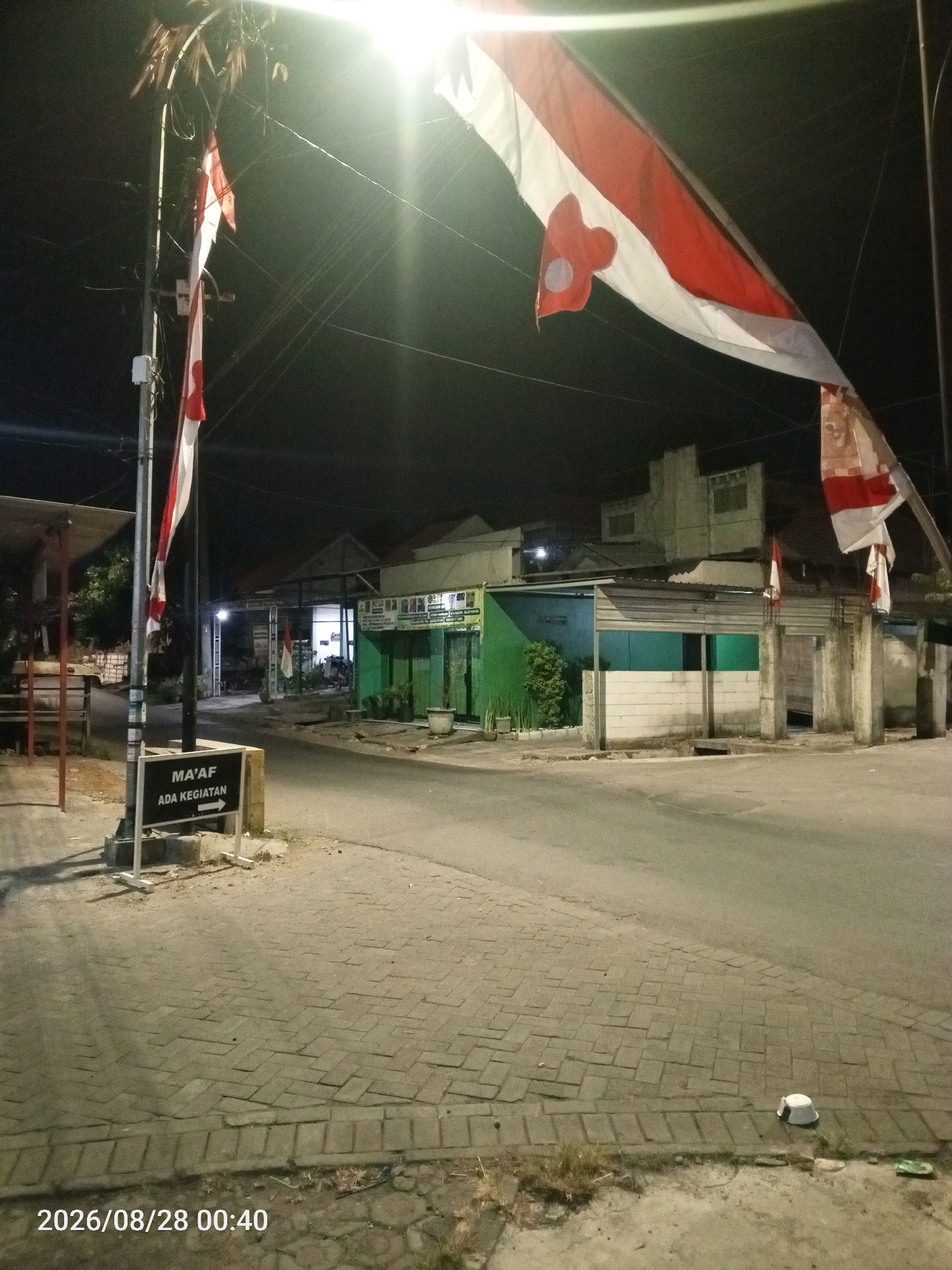 A night-time outdoor scene captures a street in Wedoroanom, Indonesia, illuminated by a bright overhead light. A pole stands on the left, adorned with red and white flags, suggesting a national celebration or event. A sign on the pole reads "MA'AF ADA KEGIATAN" (Sorry, there is an activity) with an arrow pointing to the right. The street is paved with interlocking bricks, and a road runs alongside it. Buildings with corrugated metal roofs and concrete walls line the street, some displaying storefronts with visible signage. The overall ambiance is quiet and still, with no people or vehicles present in the immediate vicinity. A discarded white cup is visible on the pavement in the lower right corner. The image is dated 2026/08/28 at 00:40.