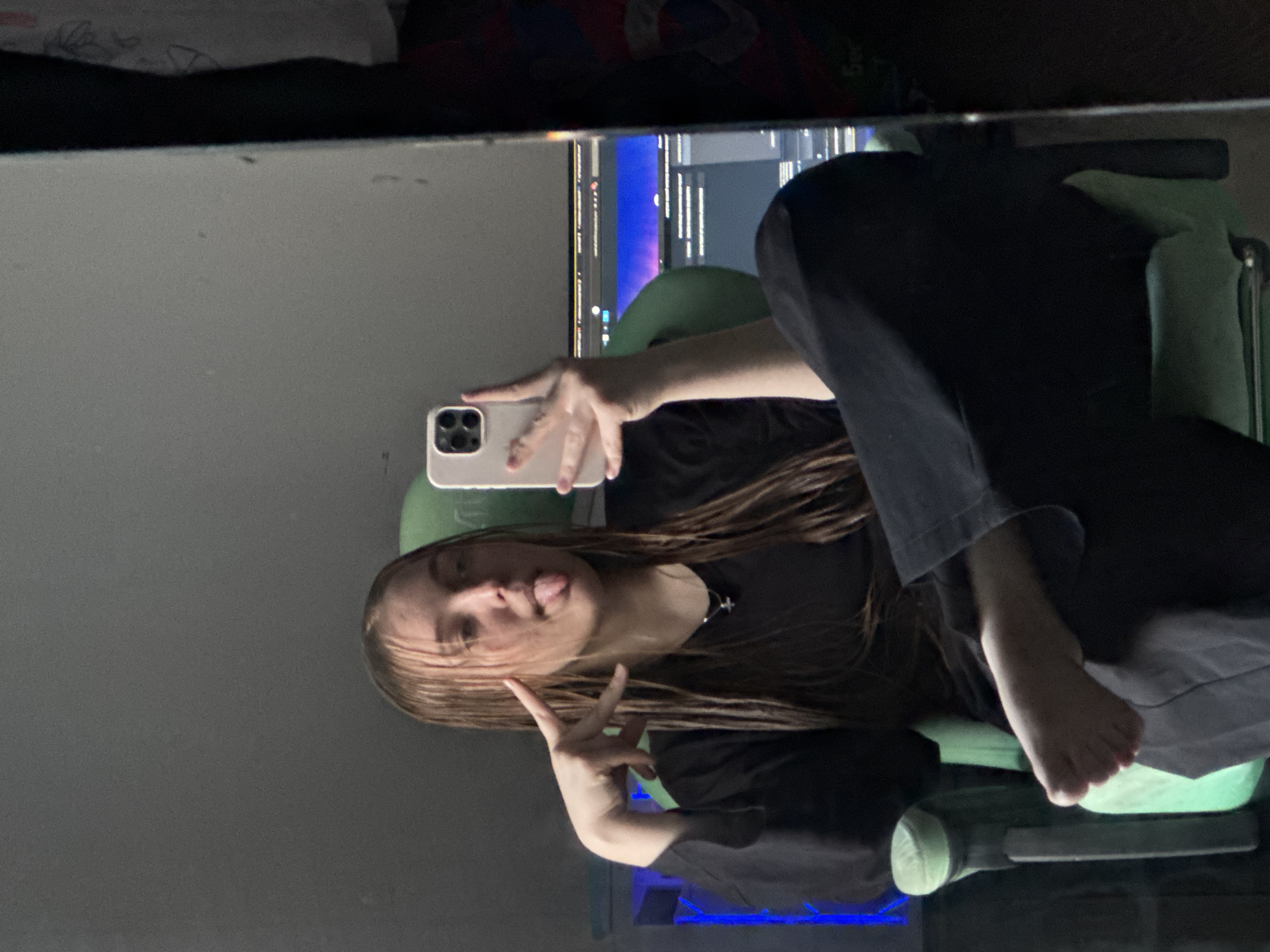 A mirror selfie captures a young individual, likely female, in an indoor setting. The person, with long, straight brown hair, is seated in a light green office chair, wearing a dark short-sleeved t-shirt and dark pants. They hold a light-colored smartphone, featuring a triple-lens camera, in their left hand, using it to take the photo. With their right hand, they make a peace sign while playfully sticking out their tongue, conveying a casual and cheerful emotion. A dark cross pendant is visible around their neck.
In the background reflection, a computer monitor displays what appears to be lines of code or text with blue and purple syntax highlighting, suggesting a desk setup for computing or gaming. The ambient lighting appears artificial. The image is oriented with a 90-degree clockwise rotation. No specific legible text is visible other than the indistinct content on the computer screen. This scene is located in Perm, Russia.