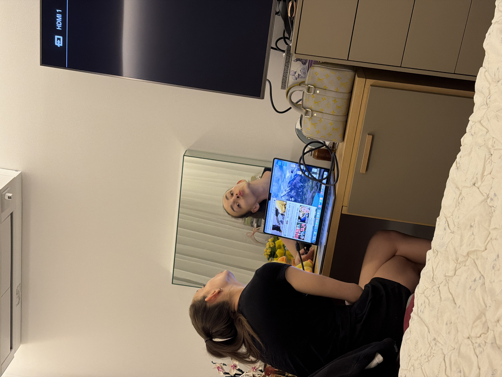 In a bedroom, a young woman with her hair in a ponytail is sitting at a desk, looking into a mirror. She is wearing a black t-shirt and shorts. Her reflection shows her making a duck face. On the desk in front of her is a laptop, open to a webpage with several images. A designer handbag with a monogram pattern is also on the desk, next to a stack of drawers. A large television screen is mounted on the wall to the right of the desk. The room appears to be modern, with neutral-colored walls and furniture. The overall impression is one of a casual, relaxed setting. The city of Sofia, Bulgaria, is not depicted in this image.