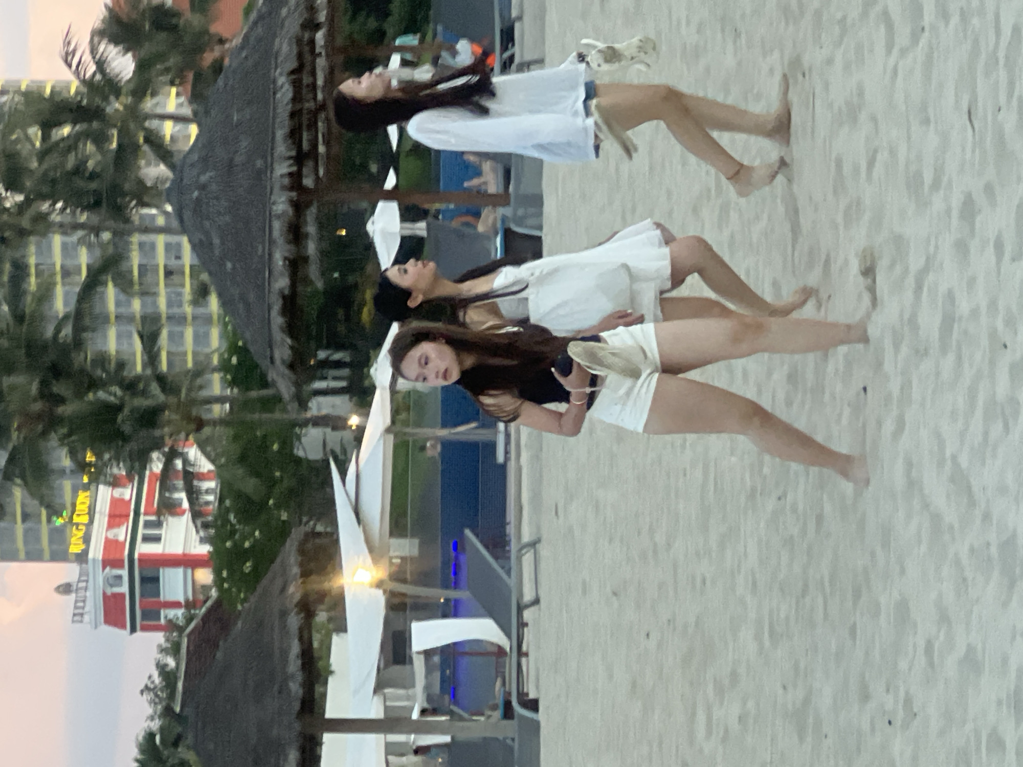 The image depicts three young women walking on a sandy beach in Da Nang, Vietnam, near a resort. In the background, a tall building with a yellow facade and a red and white building with a distinctive architectural style are visible, suggesting a hotel or resort complex. Palm trees and thatched umbrellas are scattered across the beach, indicating a tropical environment. The sky above is a soft pink and orange, suggesting it is either sunrise or sunset.
The woman in the foreground is wearing a black tank top and white shorts. She is holding a pair of flip-flops in her left hand. To her right, another woman is wearing a white dress and carrying a light-colored bag. The third woman, on the far right, is wearing a white long-sleeved top and denim shorts, and she is also holding a pair of flip-flops. All three women appear to be walking away from the camera, towards the resort in the background. The beach is mostly empty, with a few lounge chairs and umbrellas visible further back, near the water. The sand is light-colored and appears to be slightly damp in some areas.