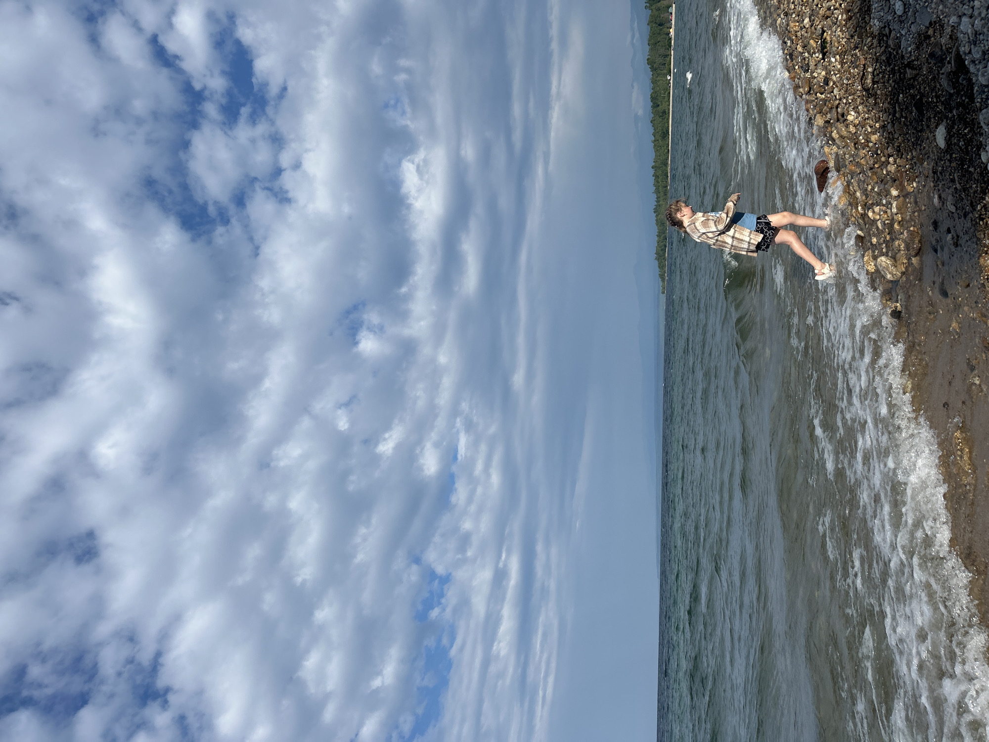 A young female with blonde hair braided down her back walks along a rocky shore. She is wearing a plaid shirt, black shorts with a white pattern, and white sandals. She is looking down at the ground as she walks. The water of a large lake is to her left, with small waves breaking on the shore. The sky above is filled with dramatic clouds. In the distance, a tree-covered shoreline is visible, with mountains beyond. The location is Tolbazikha, Russia.