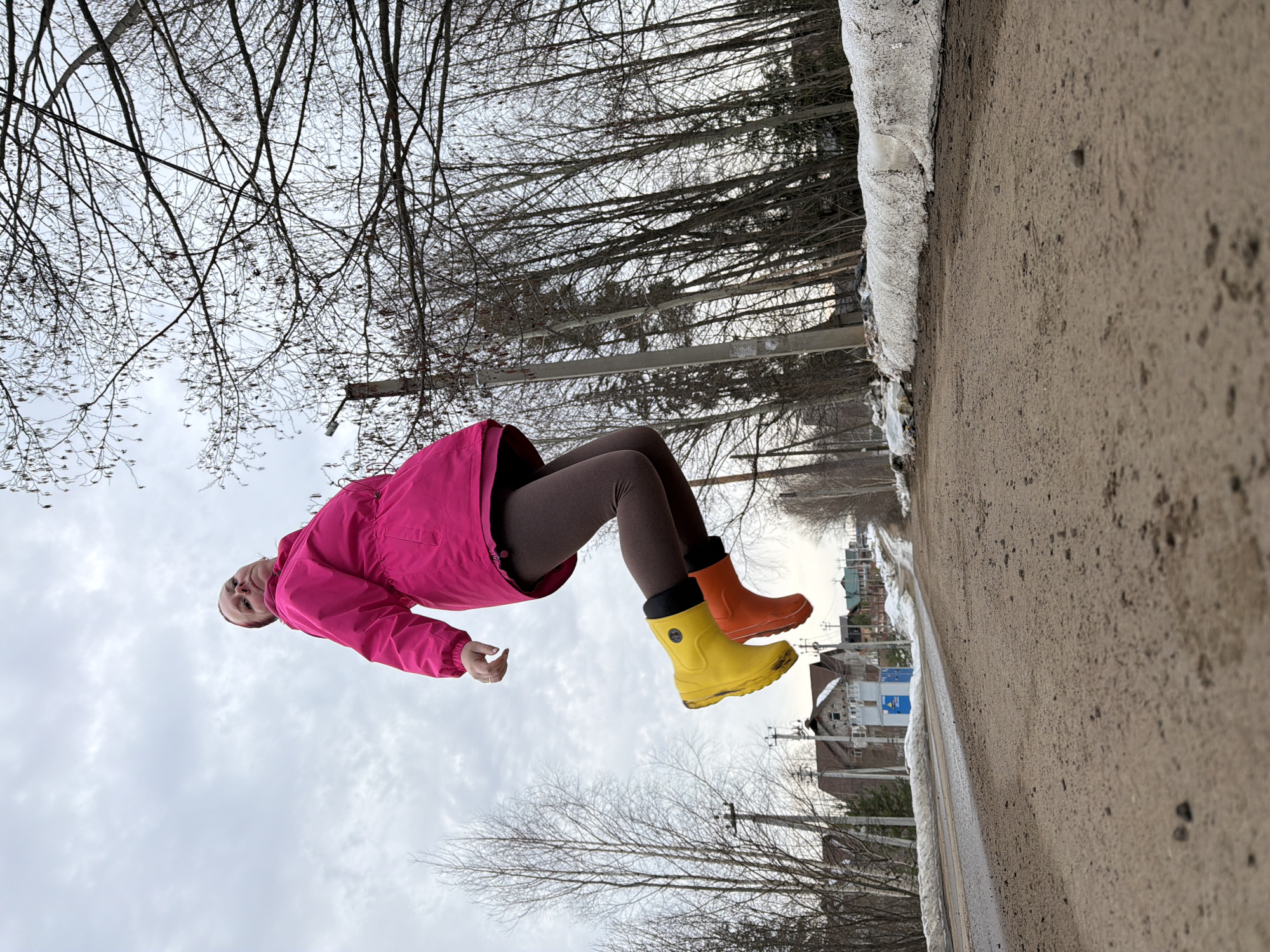 A woman in a pink jacket, brown leggings, and mismatched yellow and orange boots appears to be floating mid-air along a sidewalk. The location is likely Chaikovsky, Russia, as the buildings in the background are typical of Russian suburbs. The scene is likely a creative photographic illusion. To the right of the woman is a bare tree line, a paved path, and some snow alongside the path. To the left is a sky filled with light-colored clouds and bare trees.
