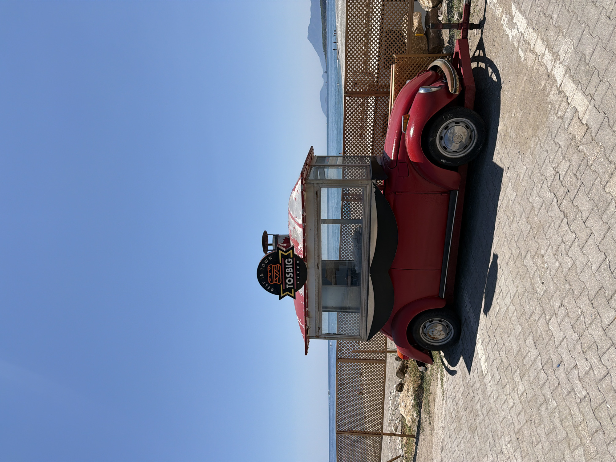 A vintage red Volkswagen Beetle, converted into a food truck named "TOSBIG," is parked on a paved area by a beach in Kemer, Turkey. The food truck has a distinctive red roof structure with glass windows, and a sign above it reads "BEST IN TOWN TOSBIG." A fence made of wooden lattice separates the food truck from the sandy beach and the calm blue sea. In the background, a few people can be seen on the beach and in the water, with a distant mountain range visible on the horizon under a clear blue sky. The overall scene is bright and sunny, suggesting a warm day.
