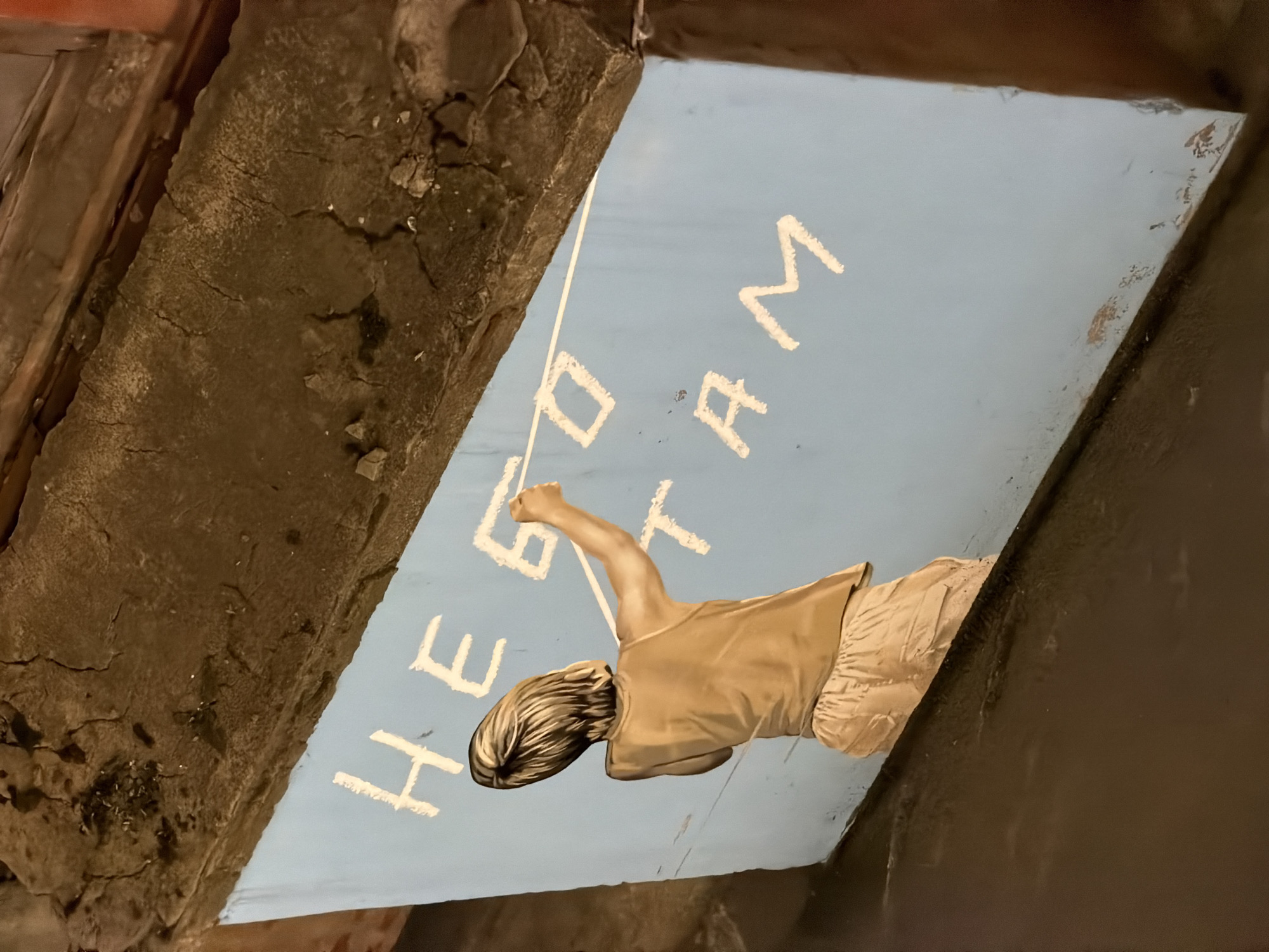 A mural in Moscow, Russia, depicts a figure reaching upwards towards the letters "HERO" in white paint on a blue surface. The mural is surrounded by aged, brown-toned architectural elements, possibly concrete or stone structures. A single white line extends from the figure's hand towards the painted letters. The figure is light-skinned, wearing a light brown top and pants. The overall perspective is looking up at the mural from below.