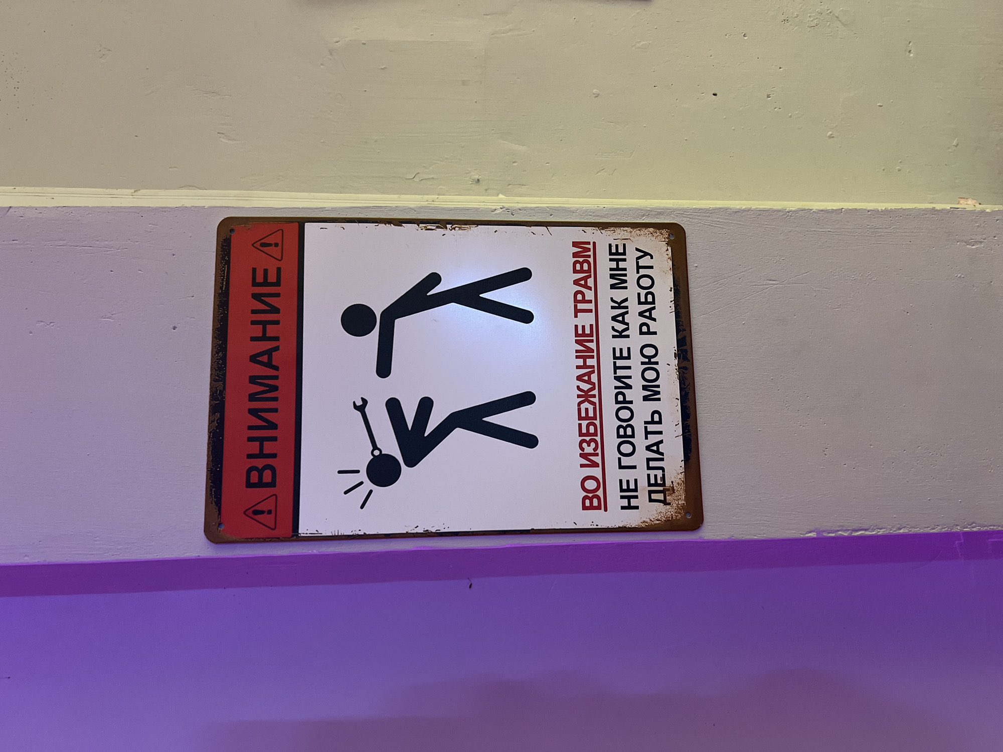 The image shows a rectangular metal sign mounted on a wall. The sign is divided into two sections: a red vertical strip on the left side, containing the word "ВНИМАНИЕ" in Cyrillic script and a warning symbol, and a white section displaying two stick figures. The top figure is standing upright, while the bottom figure is depicted lying on the ground with a wrench near its head and radiating lines indicating impact. To the right of the figures, text in Cyrillic script reads "ВО ИЗБЕЖАНИЕ ТРАВМ, НЕ ГОВОРИТЕ КАК МНЕ, ДЕЛАТЬ МОЮ РАБОТУ" (translated as "To avoid injury, don't tell me how to do my job"). The sign has a rusted border. The wall behind the sign is off-white and a horizontal purple bar is running near the bottom of the image. No evidence or indication in the image suggests the location is Bắc Nha Trang, Vietnam.